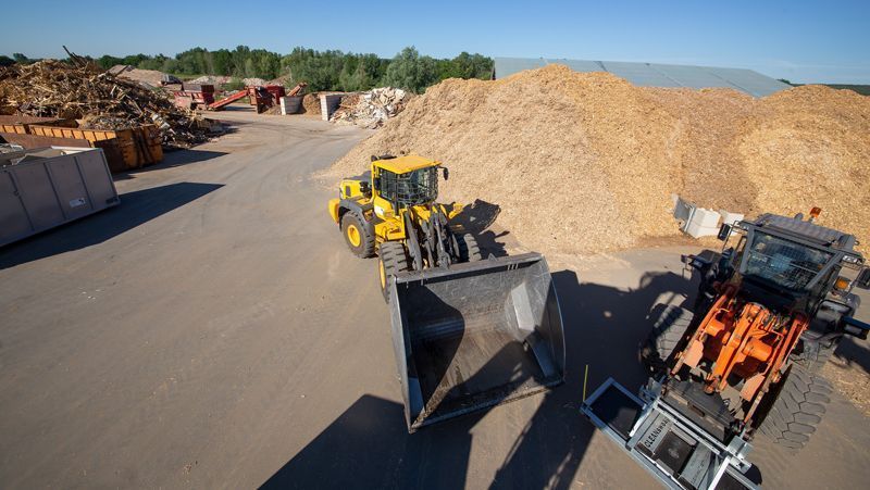 Zwei gelbe Frontlader in einem Holzverarbeitungshof mit Stapeln aus Holzspänen und Holzstämmen.