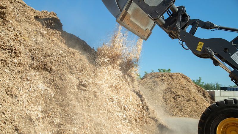 Frontlader kippt Holzspäne unter blauem Himmel auf einen großen Haufen.
