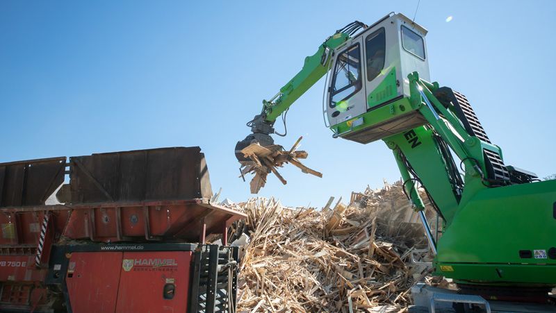 Grüner Baggerarm legt Holzstücke in eine rote Schleifmaschine, im Freien.