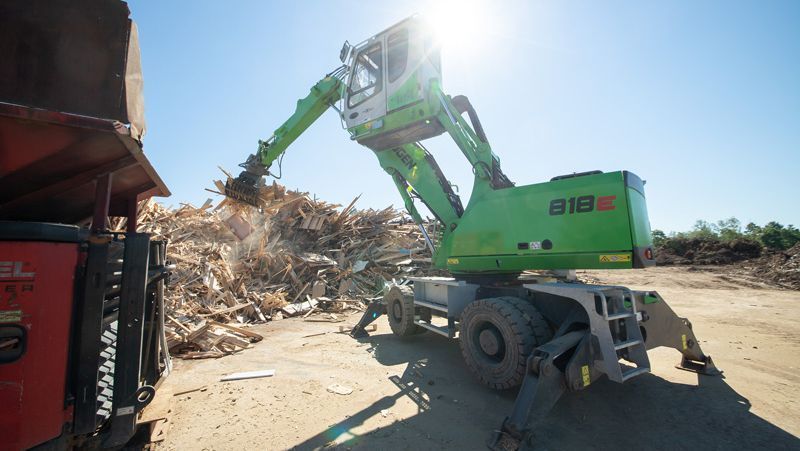Grüne Industriemaschine, die an einem sonnigen Tag Holzabfälle auf einen roten LKW lädt.