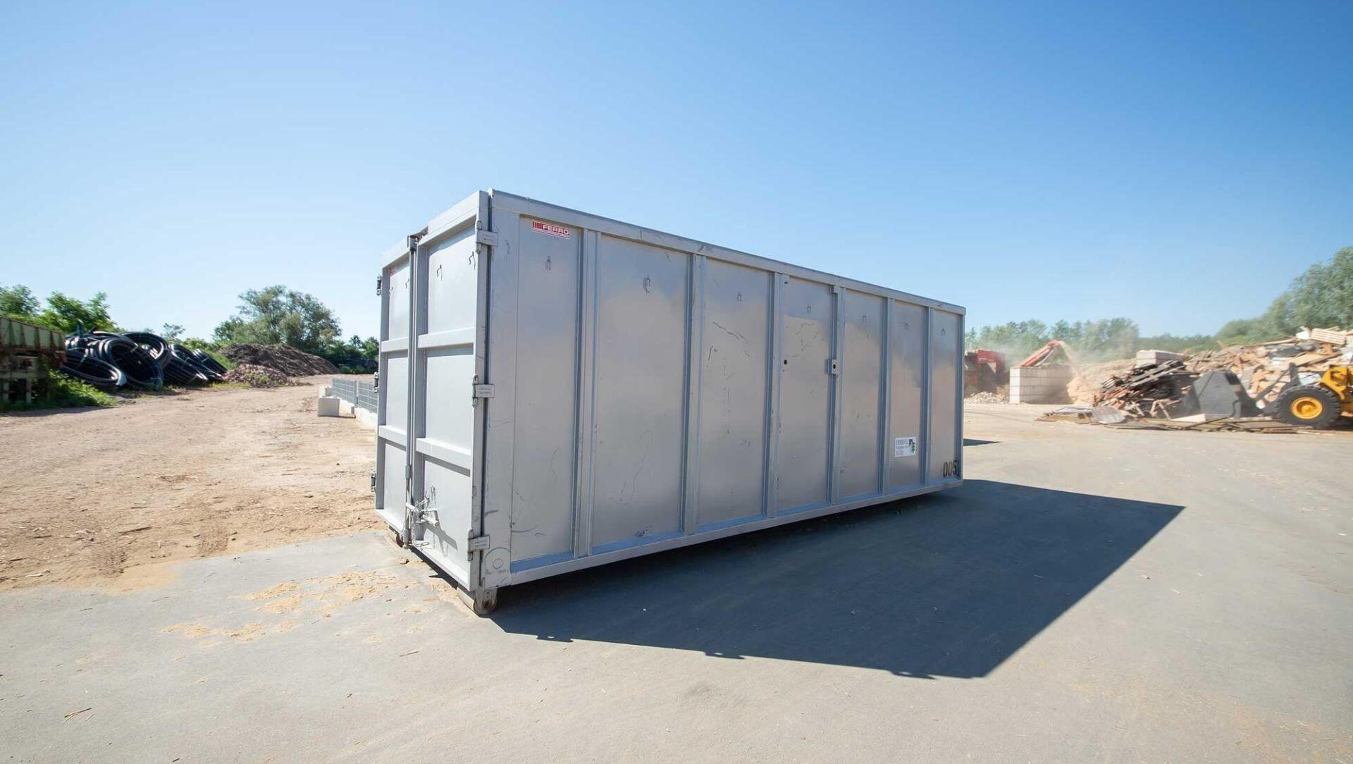 Grauer Metallmüllcontainer auf einer Betonoberfläche, mit Schutt und Maschinen im Hintergrund unter einem blauen Himmel.
