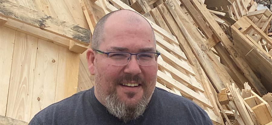 Mann mit Brille lächelt, im Hintergrund ein Holzstapel.