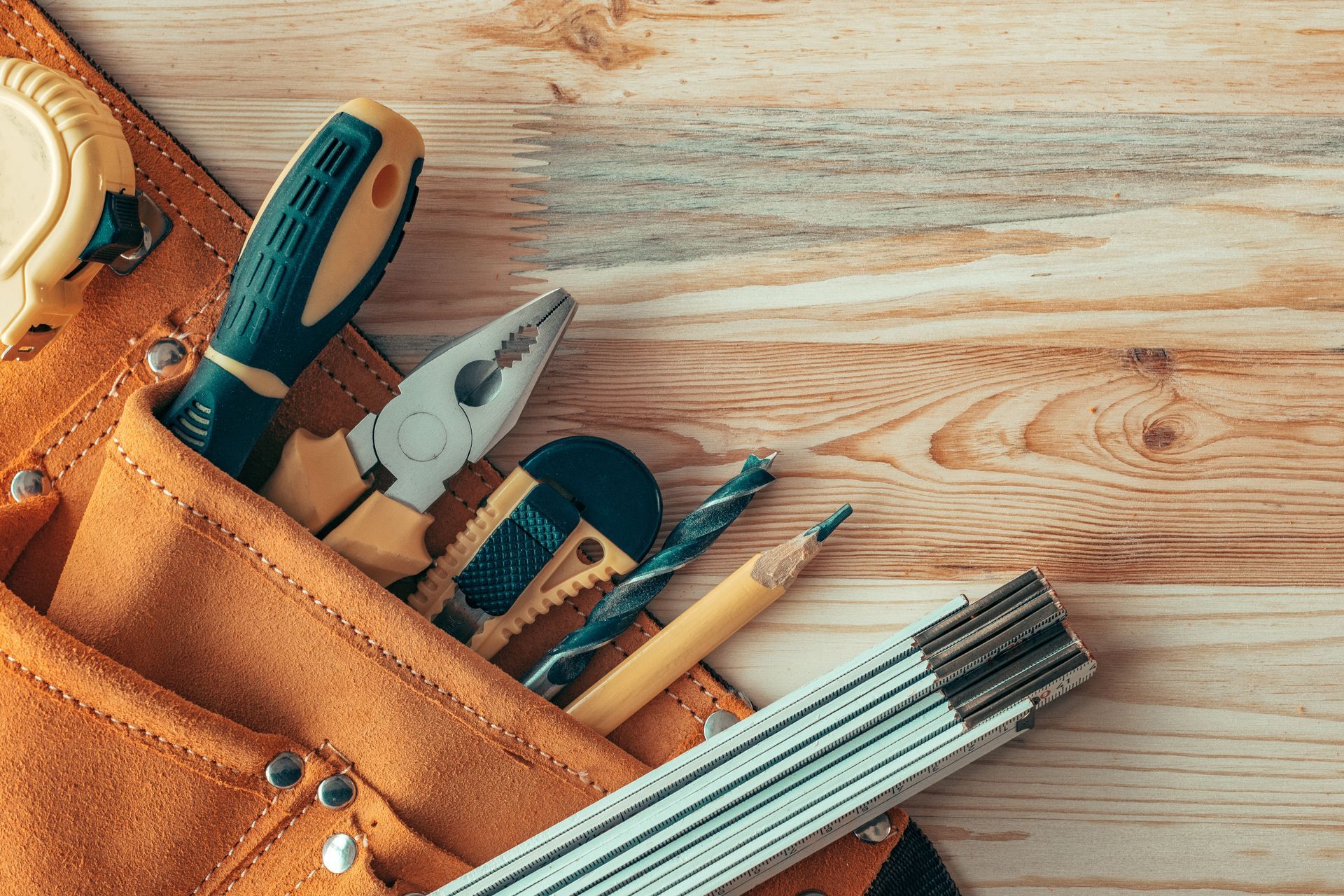 Une ceinture à outils contenant un tournevis, une pince, un couteau utilitaire, un foret, un crayon et une règle pliante, posée sur une surface en bois.