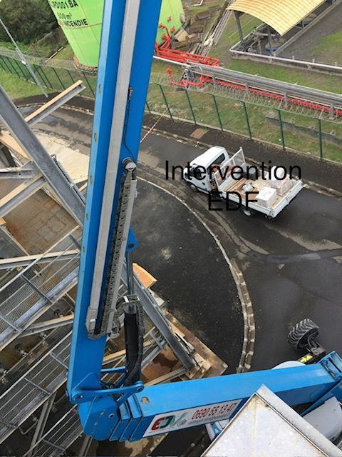 Un ascenseur de chantier bleu longe une voie ferrée encore humide, avec un véhicule utilitaire blanc en dessous et une barrière de sécurité en arrière-plan.
