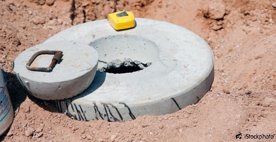 Plaque d'égout en béton avec trappe ouverte dans la terre, surmontée d'un mètre ruban jaune et d'une petite poignée noire.