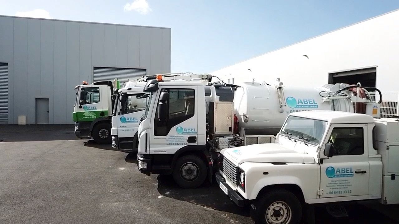 Trois camions utilitaires blancs sont garés à côté d'un entrepôt, avec un camion vert en arrière-plan.