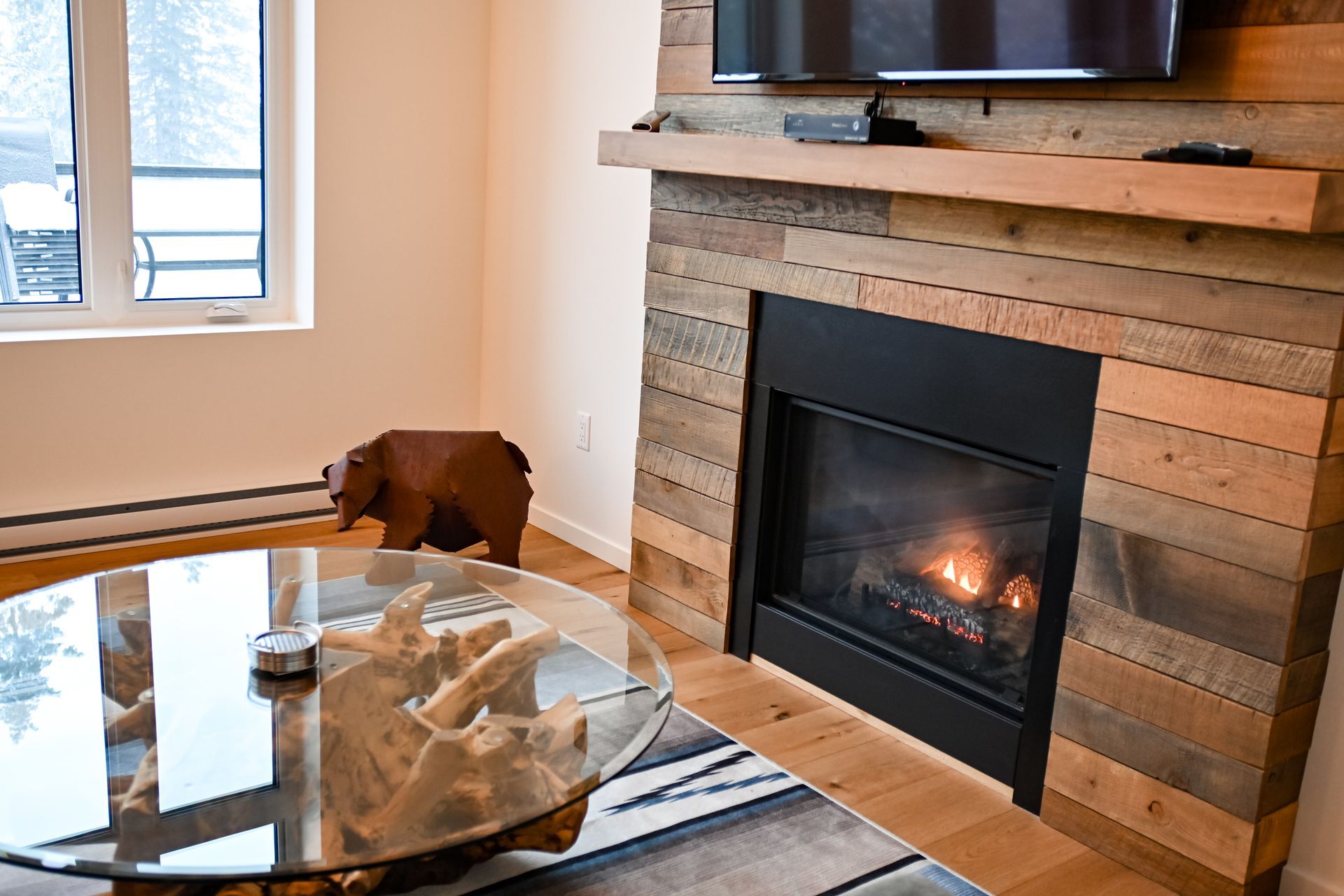 A brown dog stands by a glass coffee table near a lit fireplace in a bright living room.