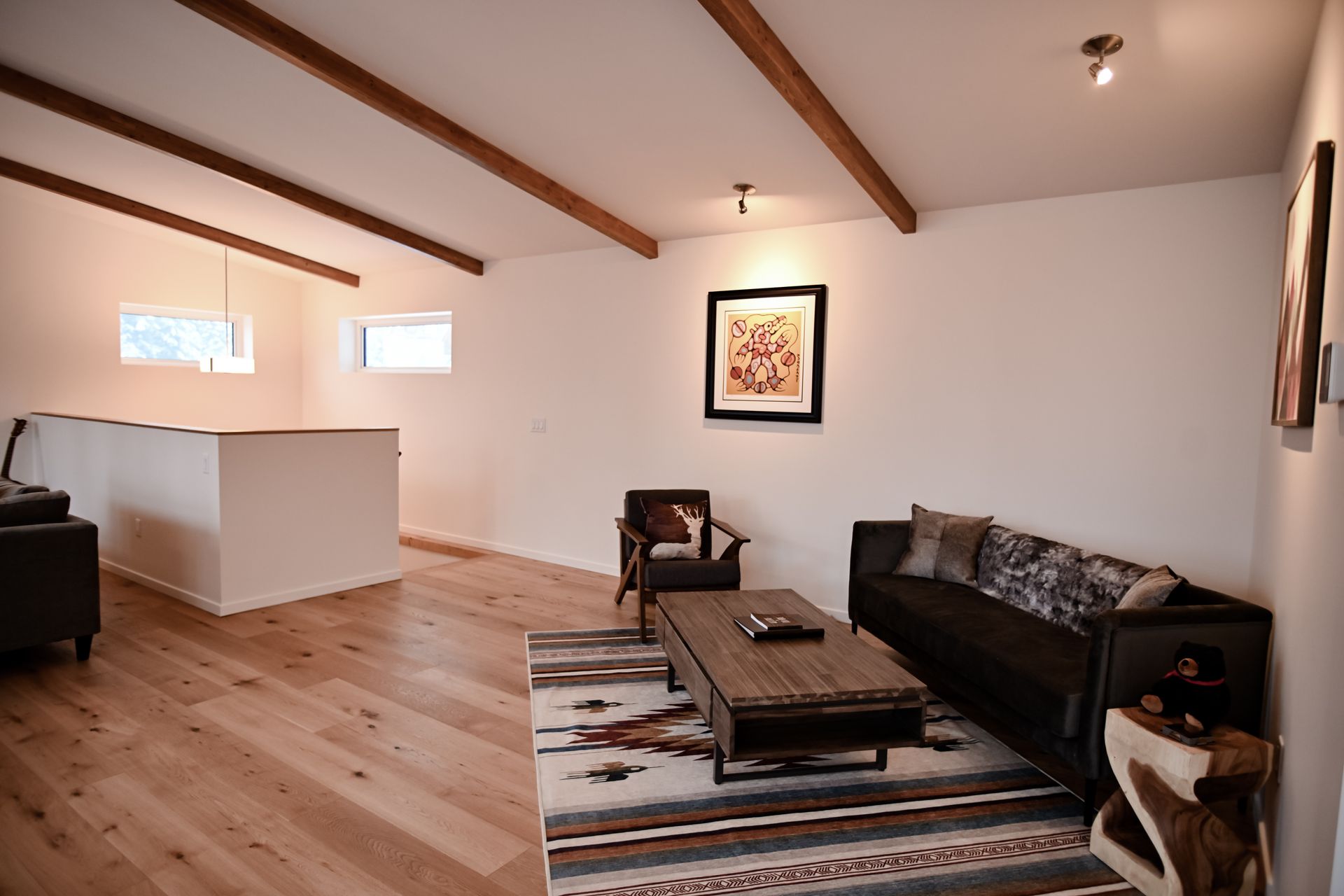 Modern living room with wood beams, black sofa, patterned rug, coffee table, and open kitchen counter.