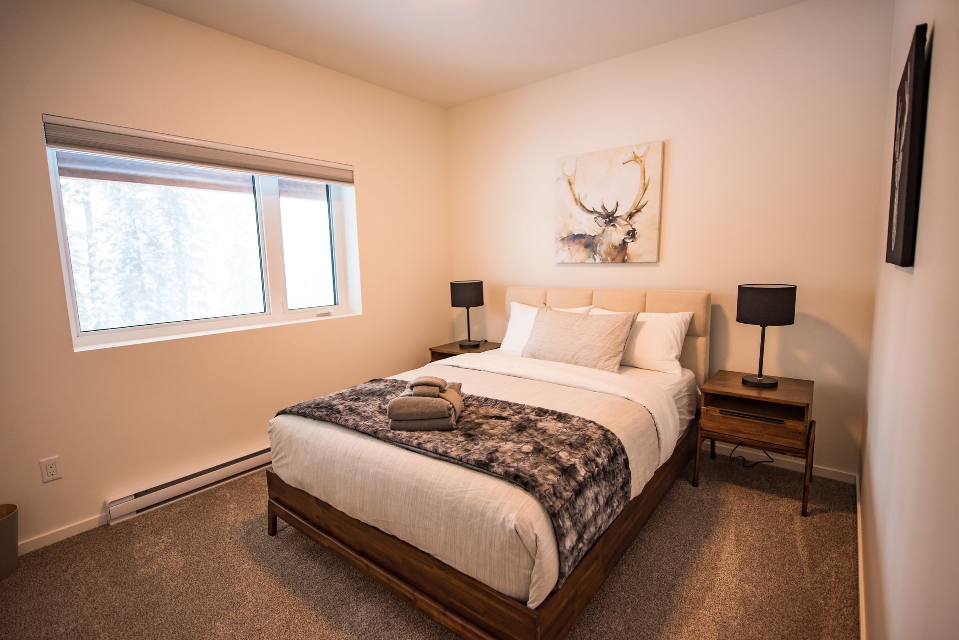 Neatly made bedroom with a wooden bed, two bedside lamps, and a window letting in daylight