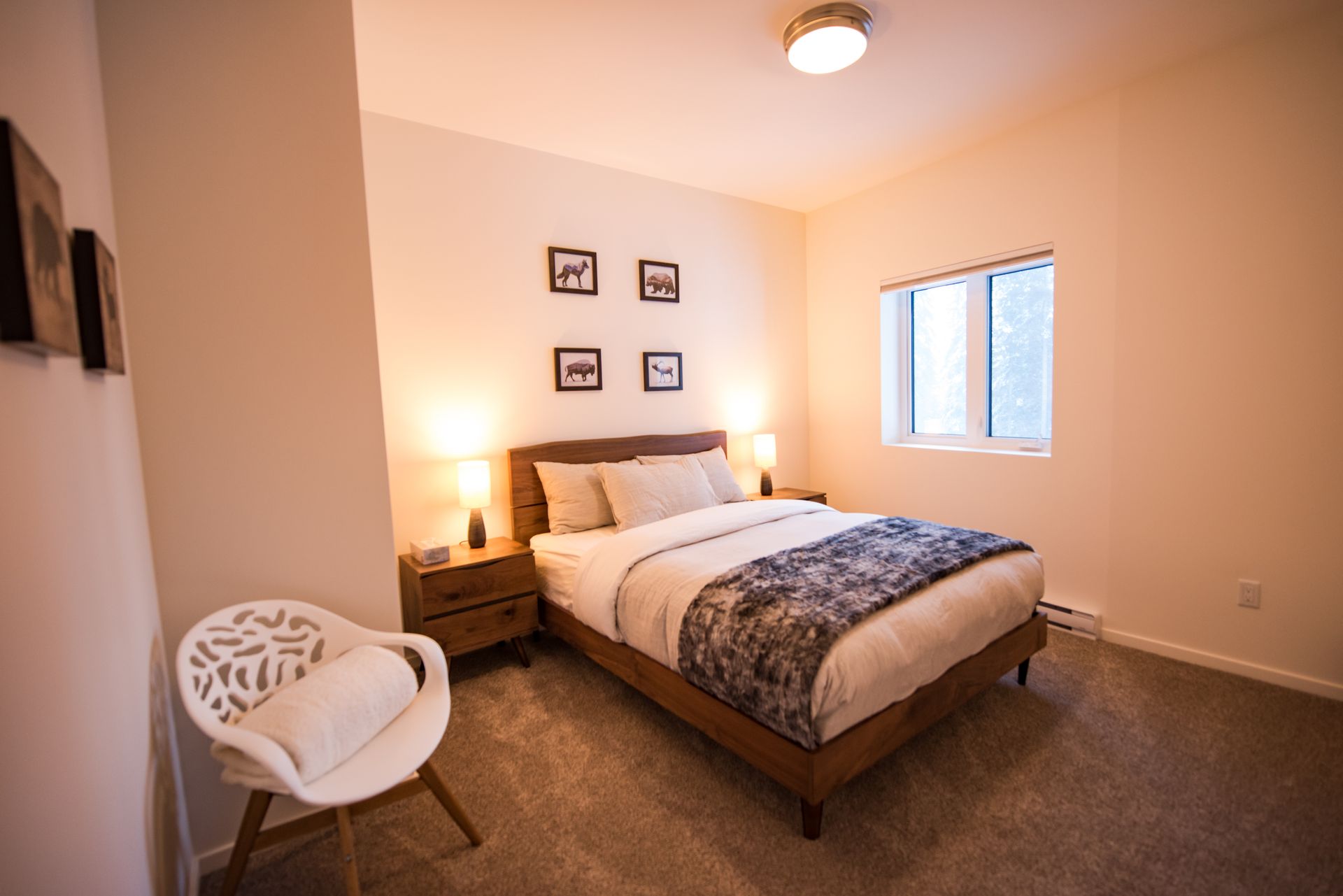 Cozy bedroom with a made bed, bedside lamps, framed wall art, and a white chair by the window
