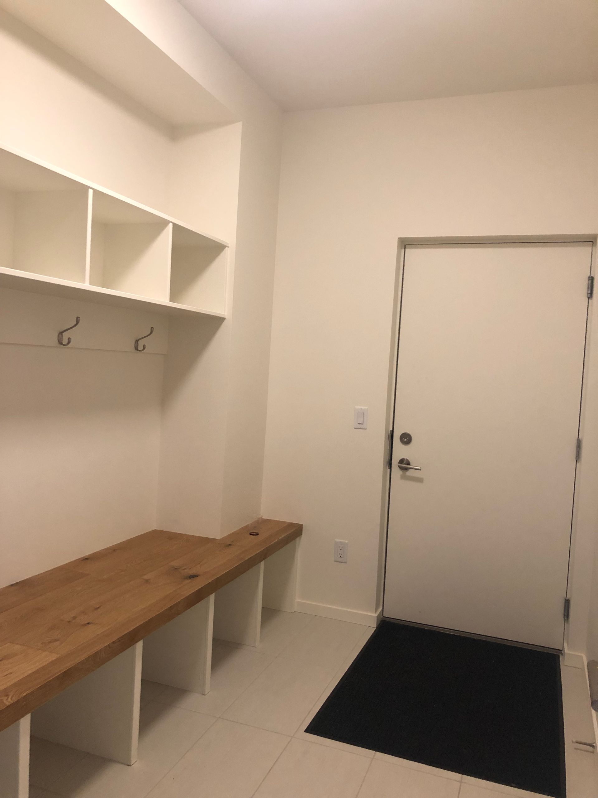 Small entryway with bench, wall hooks, overhead shelves, and a white door with a black mat.