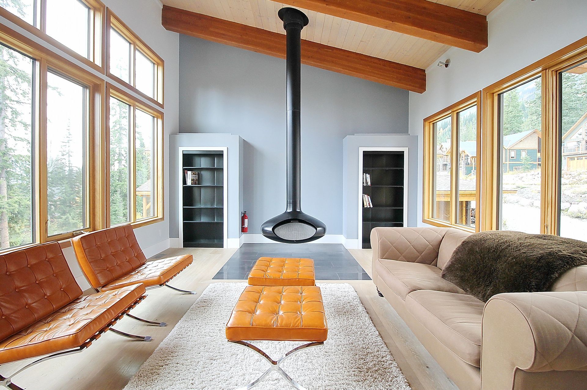 Bright modern living room with wood-beam ceiling, fireplace, sofa, wooden lounge chair, and orange ottoman