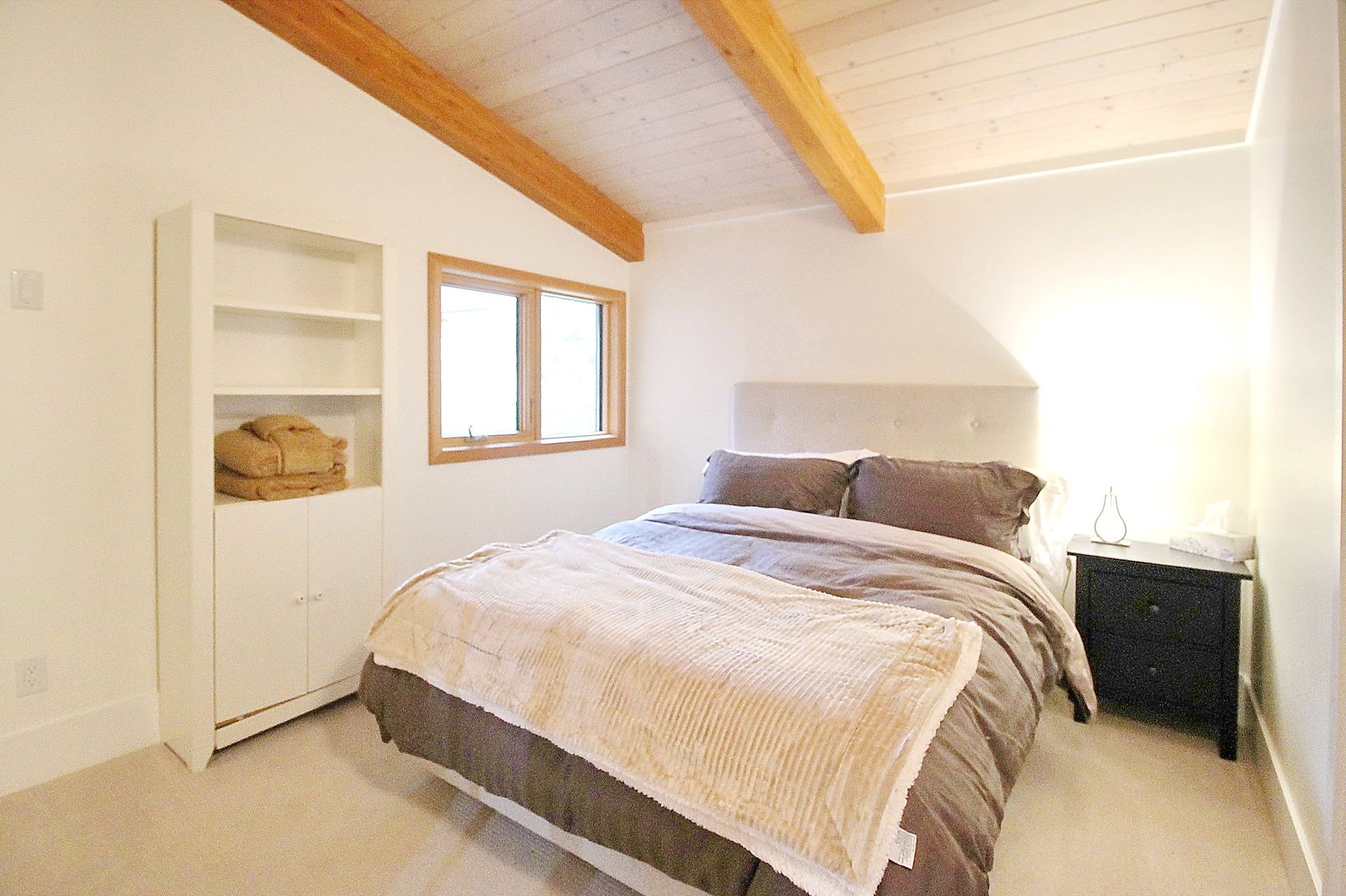 Bright bedroom with a bed, wooden ceiling beams, white walls, and a small window.