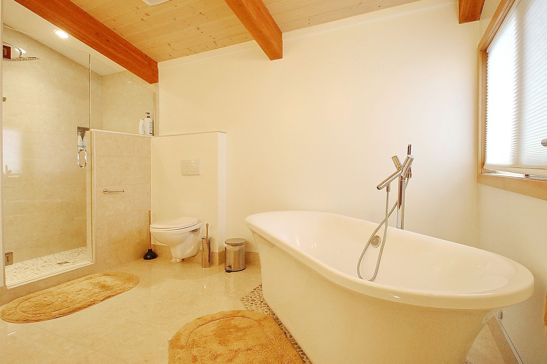 Bright bathroom with a freestanding tub, glass shower, toilet, and wood-beamed ceiling