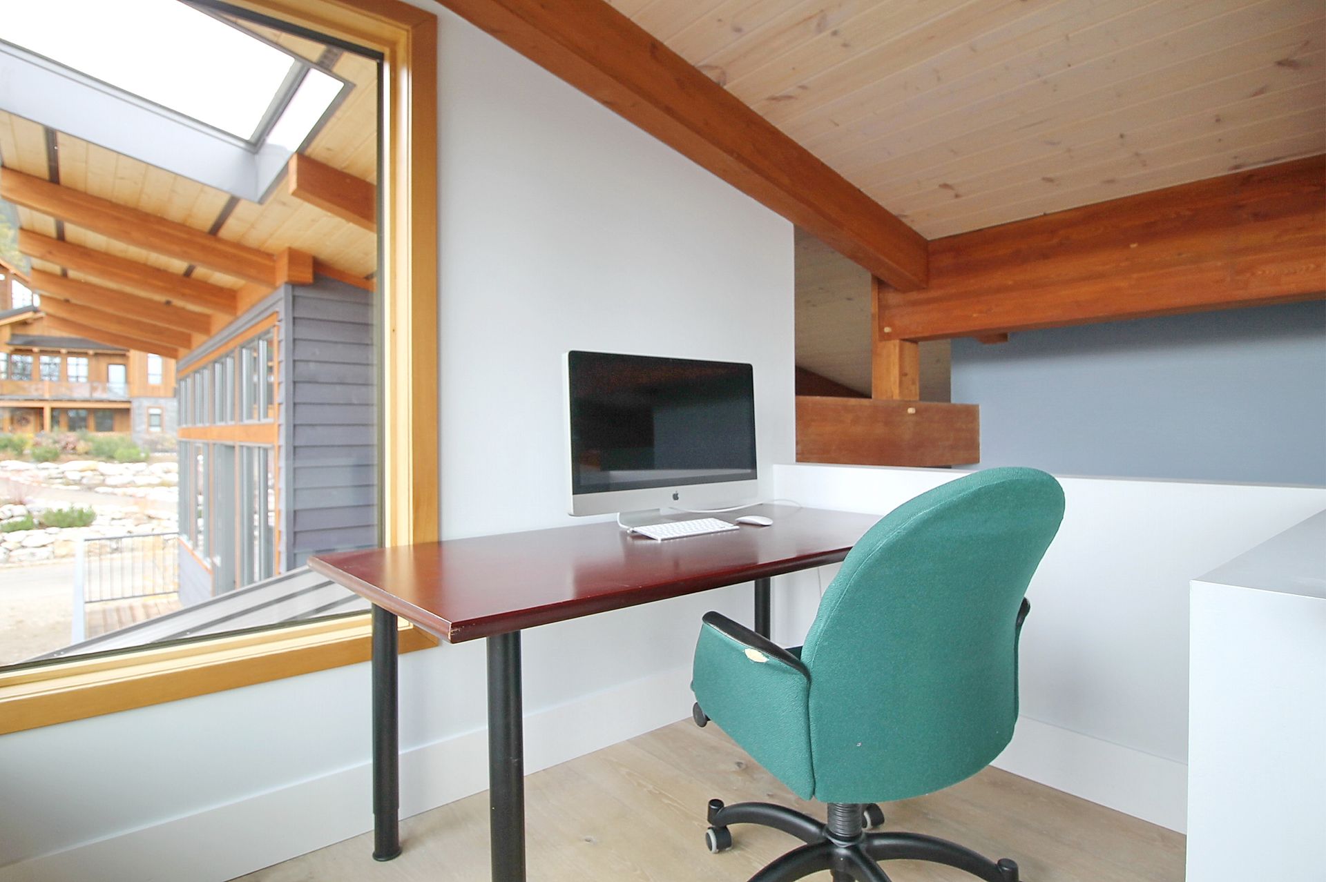 Modern loft office desk with computer, teal chair, and slanted wood ceiling by a large window