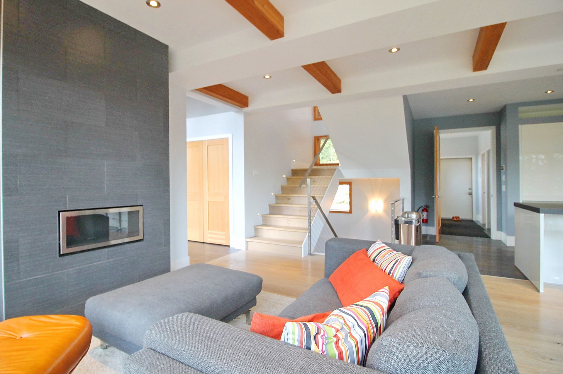Modern open-plan living room with gray sofa, orange cushions, fireplace, staircase, and kitchen in the background