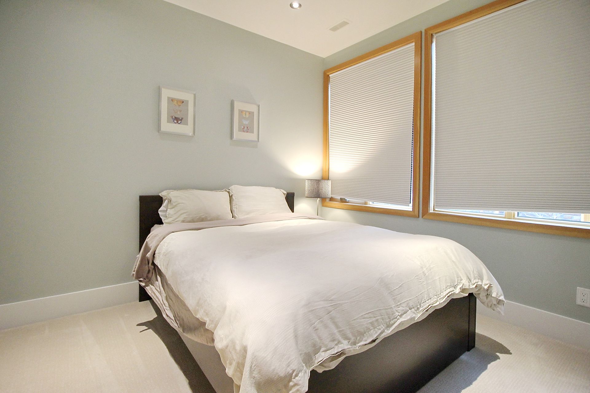 Bright bedroom with a made bed, two framed pictures, and sunlight through large windows with blinds