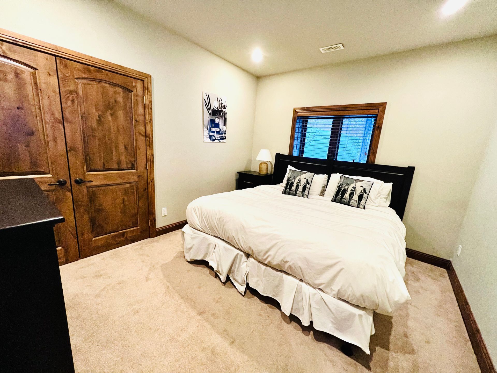 Bright bedroom with a white bed, wooden closet, and window with blue blinds on beige carpet.