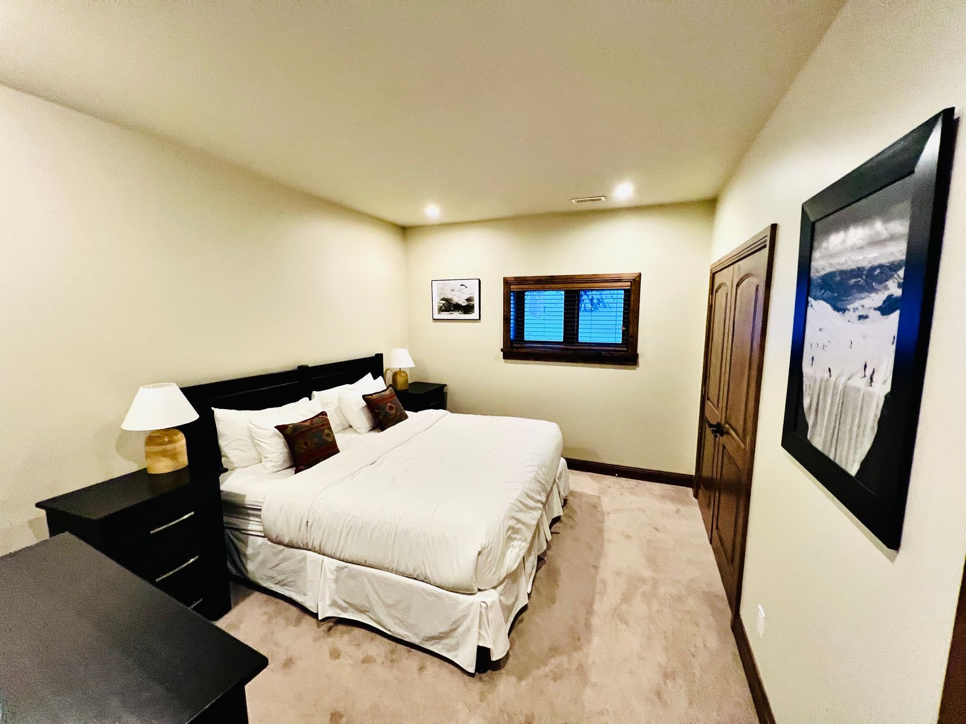 Narrow bedroom with a bed, dark nightstands, beige carpet, and framed wall art.