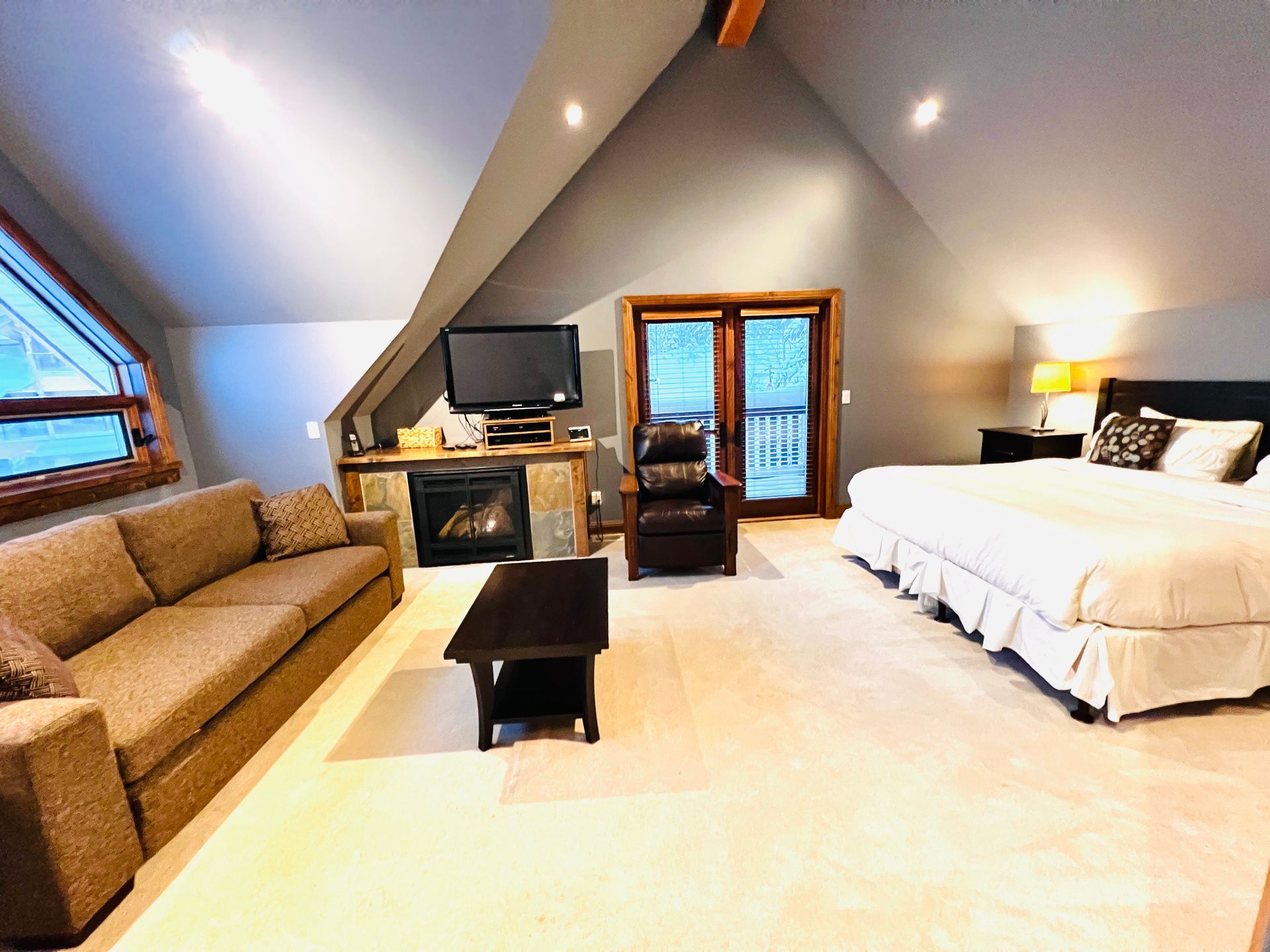 Attic bedroom with sloped ceiling, white bed, brown sofa, and TV by the fireplace