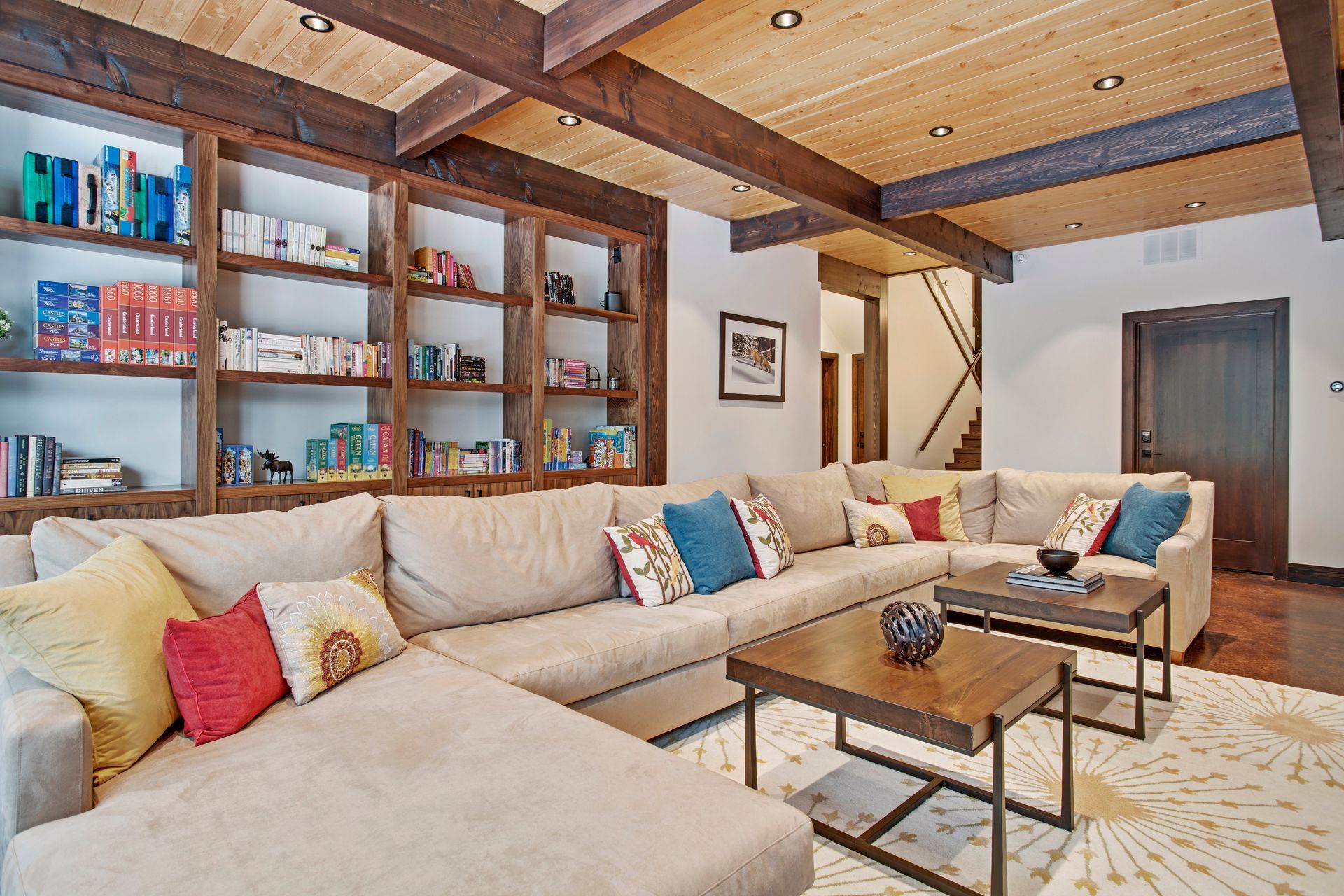 Cozy living room with beige sectional sofa, wood beams, bookshelves, and coffee tables