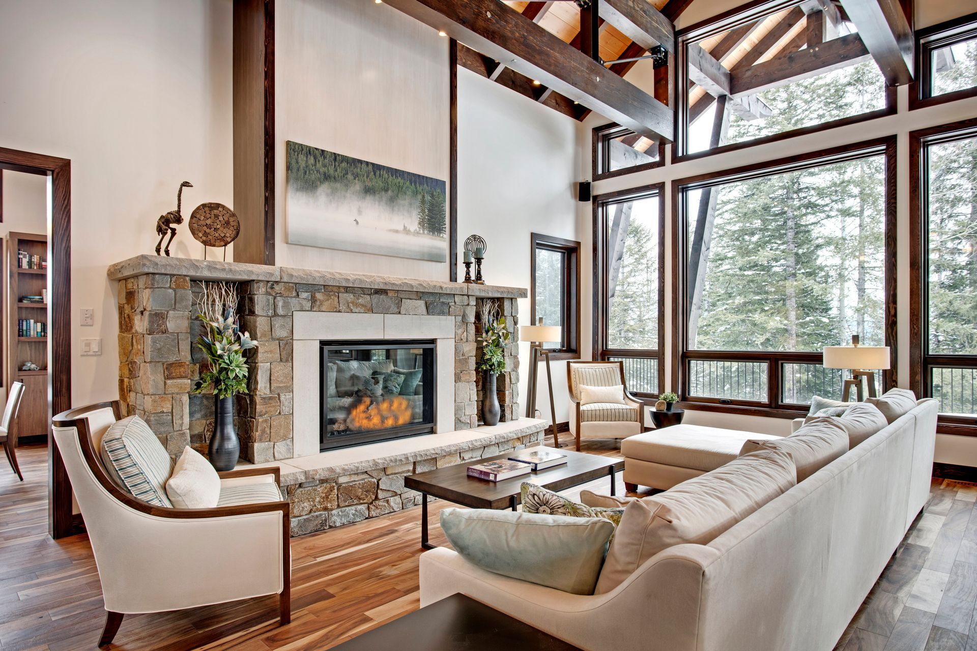 Bright living room with stone fireplace, white sectional sofa, and large windows overlooking trees