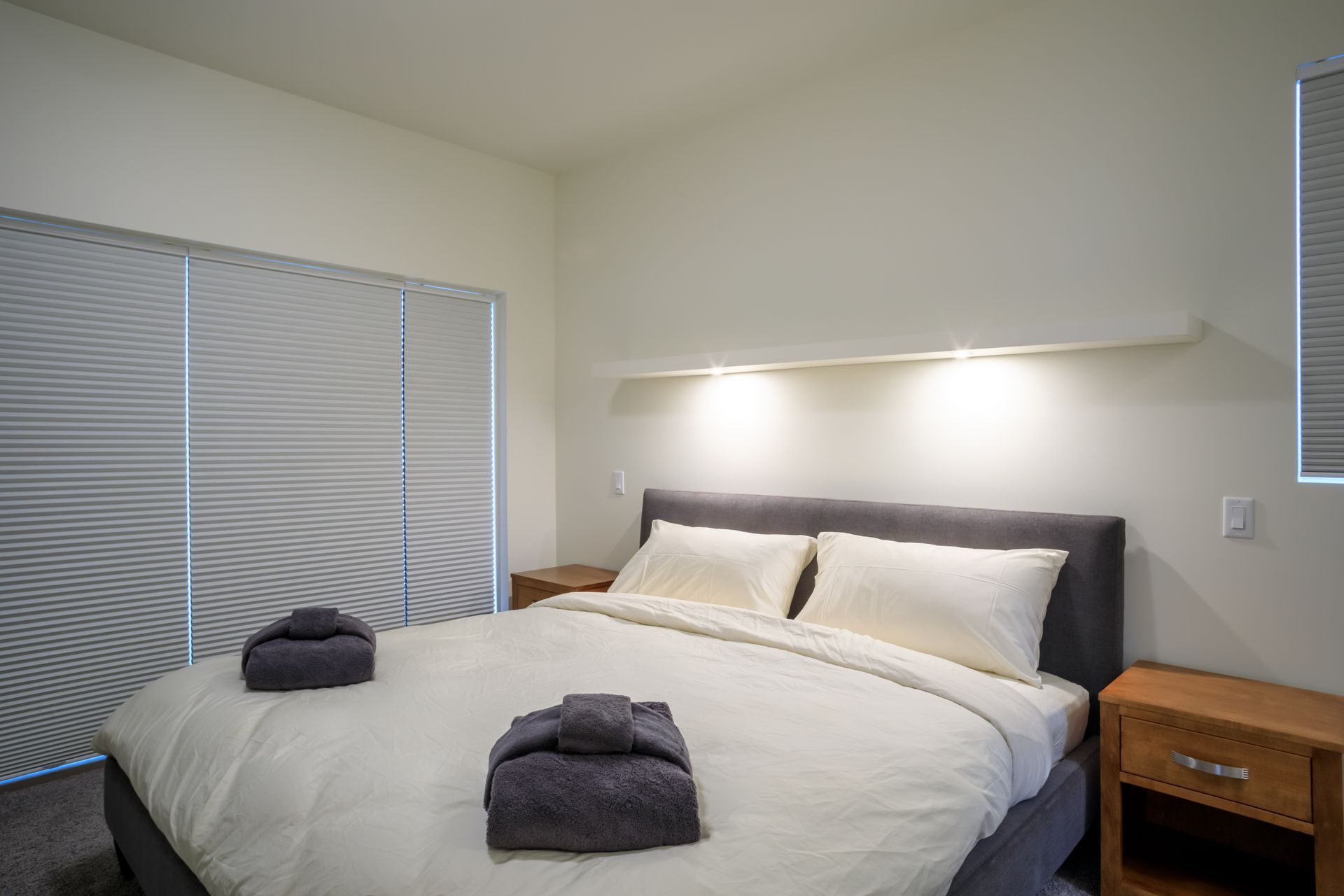 Modern bedroom with a neatly made bed, two folded towels, bedside tables, and soft wall lighting