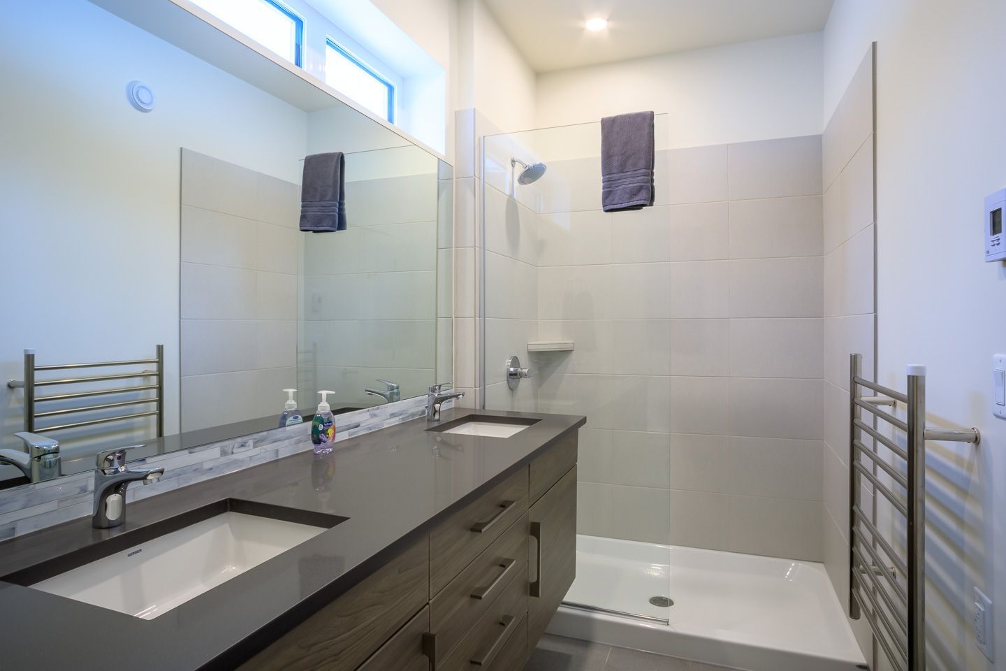 Modern bathroom with double sink vanity, large mirror, glass shower, and light gray tile.