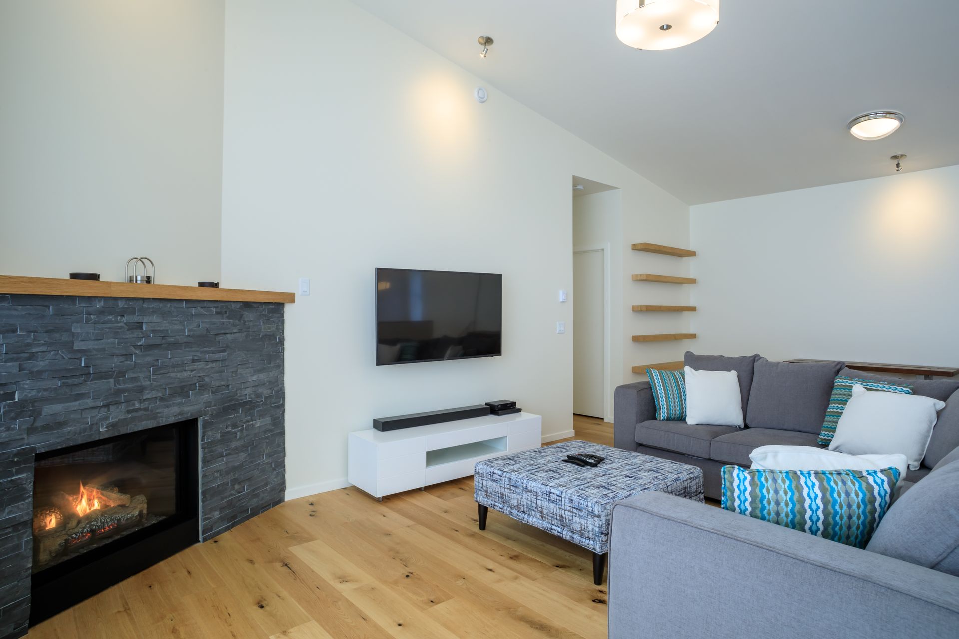 Modern living room with stone fireplace, wall-mounted TV, gray sofa, and open wooden shelves