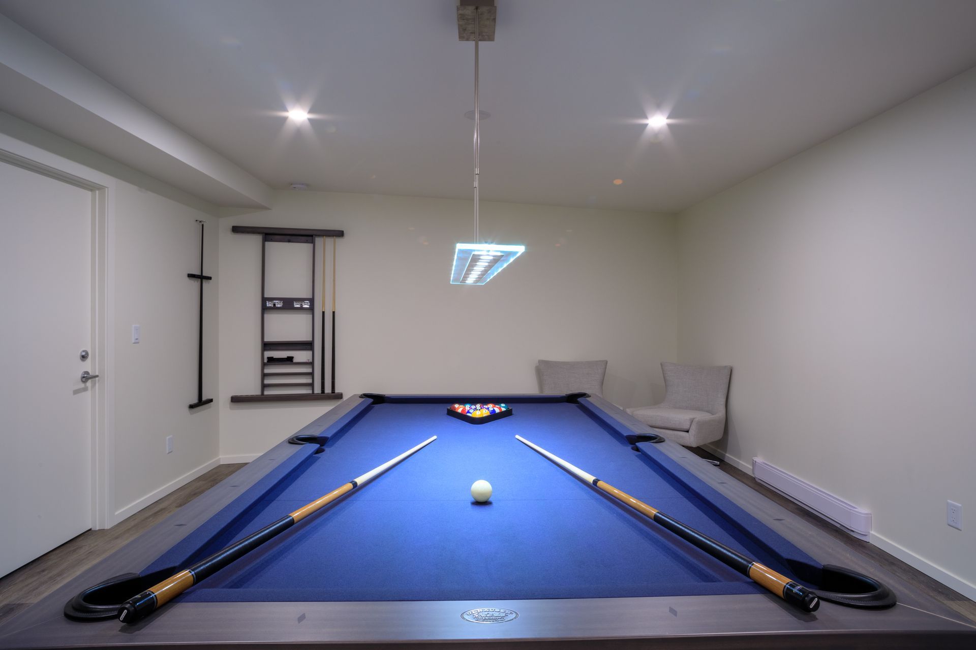 Blue-felt pool table in a white basement game room with pool cues and a small ball.