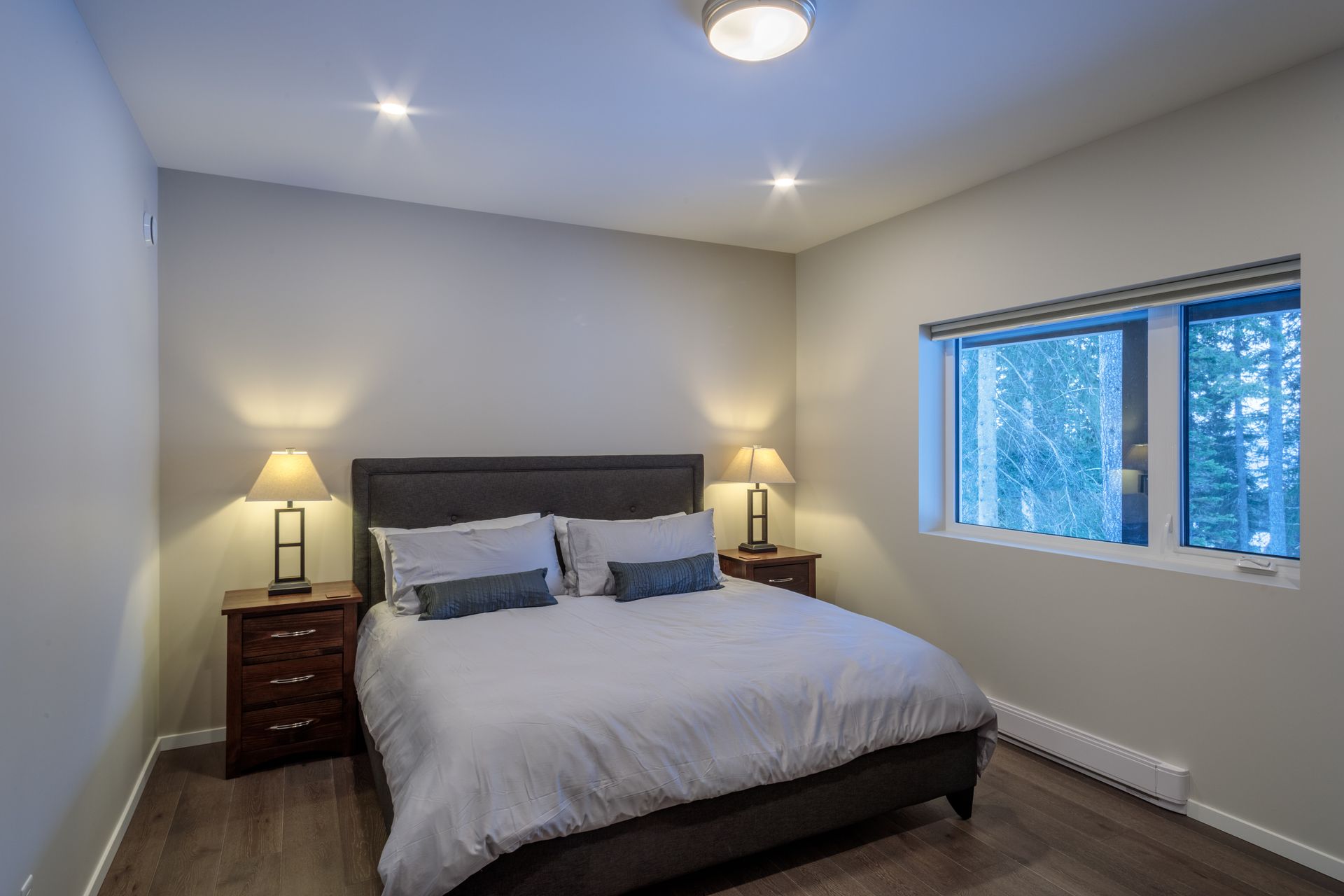 Bright bedroom with a made bed, two bedside lamps, and a window showing trees outside.