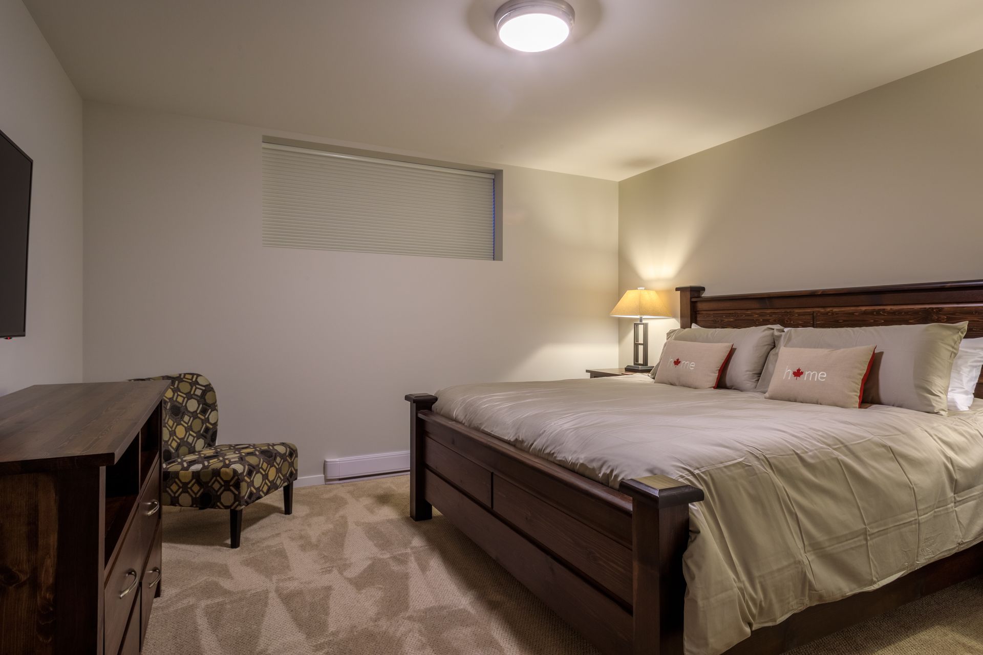 Bedroom with a large bed, lamp, chair, and dresser in a softly lit neutral-toned room