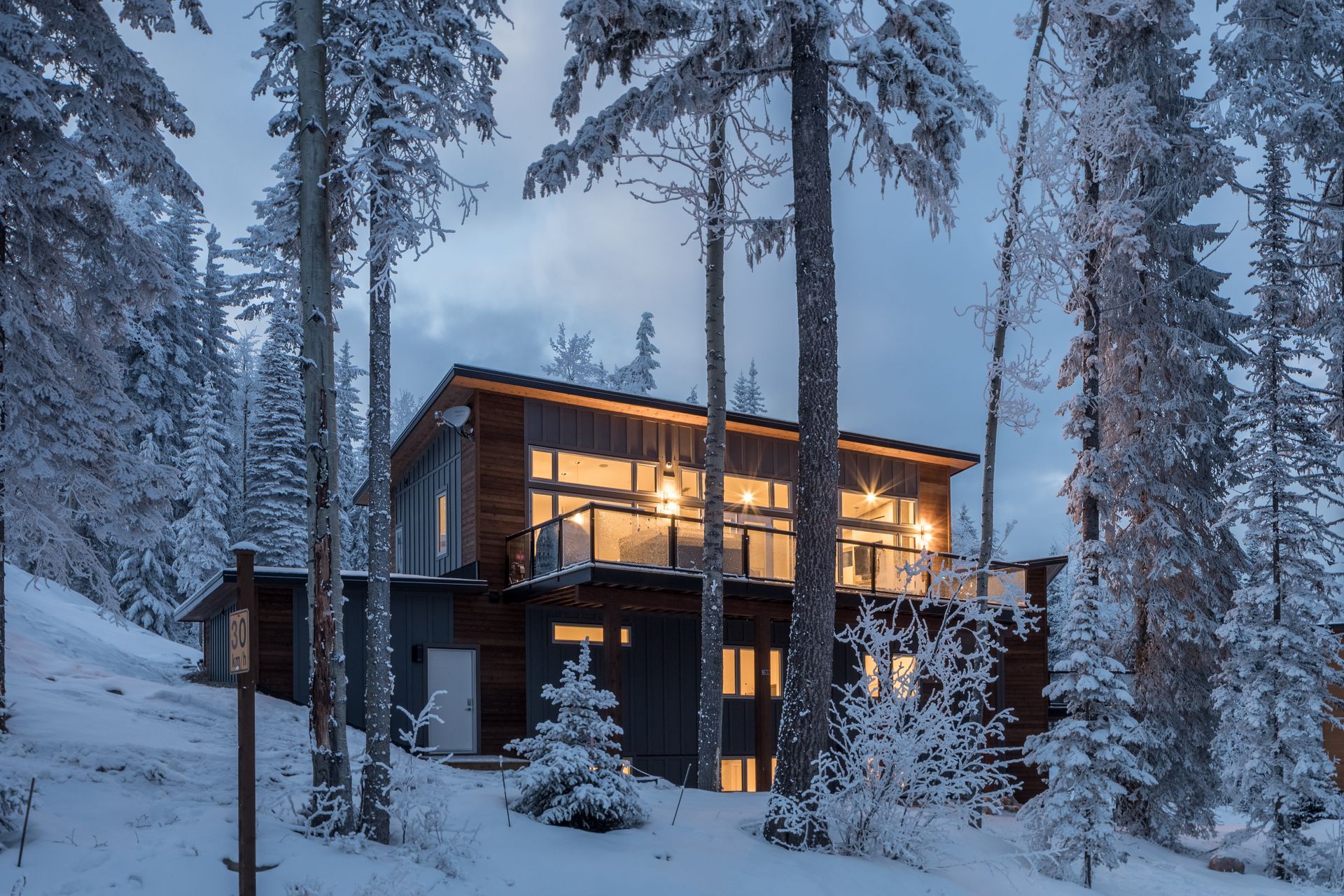 Modern house glowing warmly in a snowy pine forest at dusk
