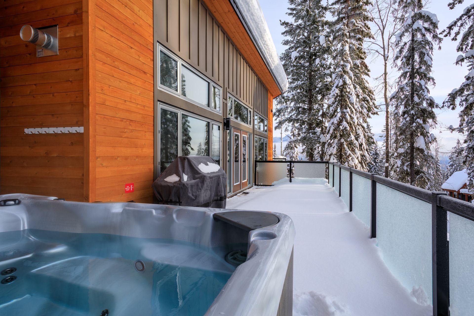 Snowy balcony with a hot tub beside a modern wooden cabin and glass railing overlooking trees