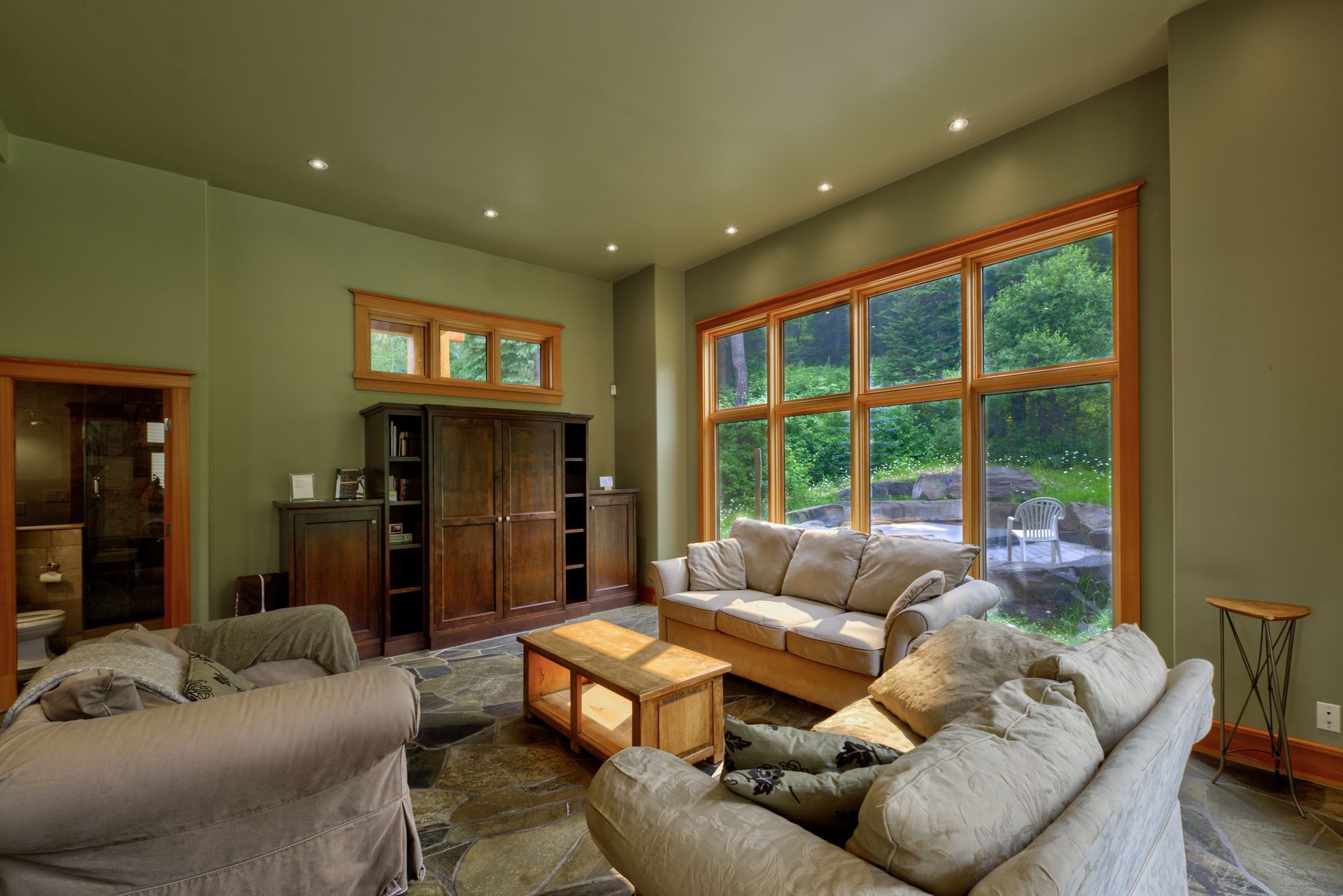 Cozy living room with large windows, sofas, coffee table, and wooden cabinets overlooking green trees