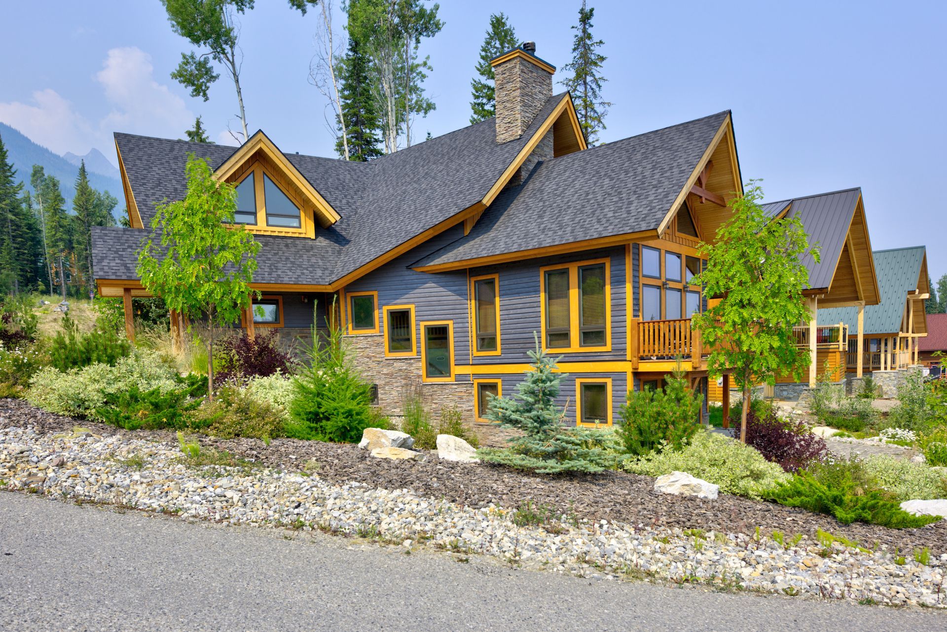 Lodge-style house with dark roof and yellow trim, set among trees and landscaping on a gravel road