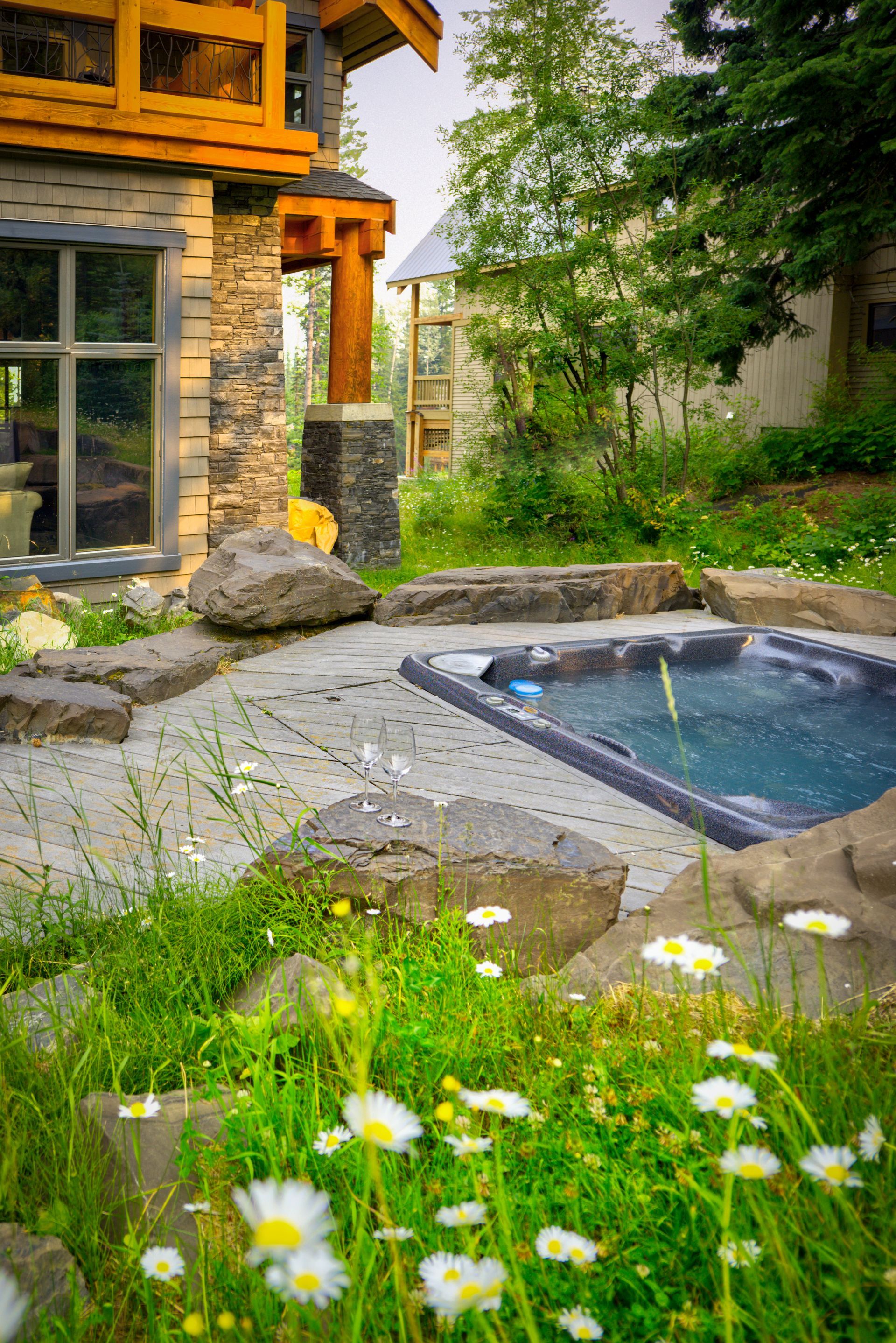 Stone patio with a small hot tub beside a house, surrounded by greenery and white wildflowers.