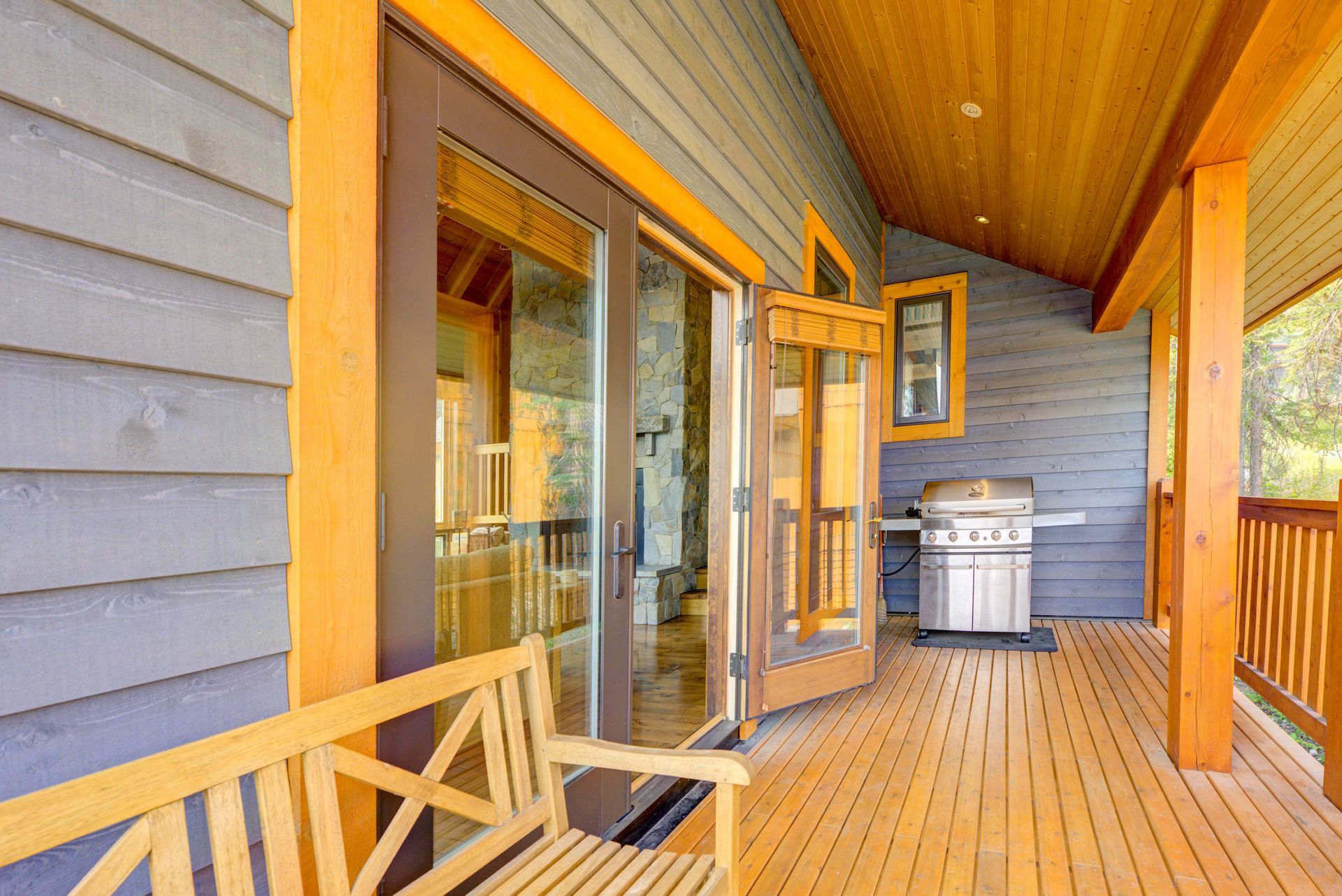 Covered wooden porch with glass doors, blue siding, and a silver grill on a sunlit deck