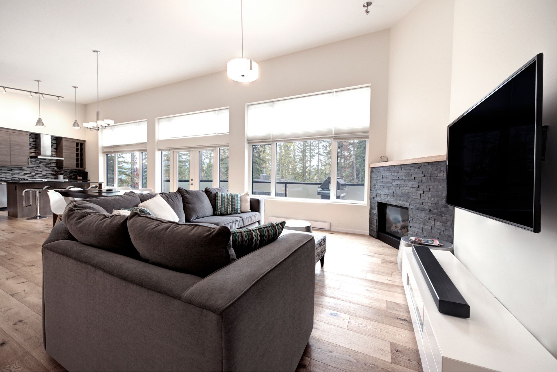 Bright open living room with gray sectional sofa, wall-mounted TV, and large sunlit windows