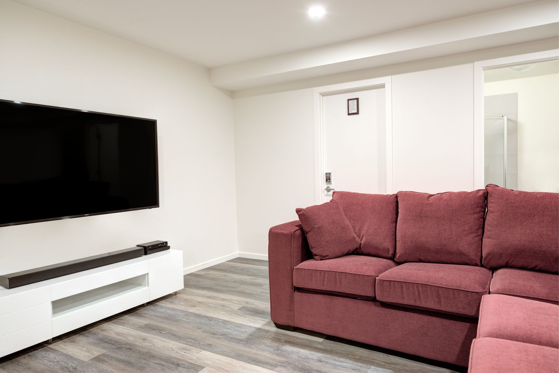 Modern living room with wall-mounted TV, white console, and maroon sectional sofa beside a bright doorway.