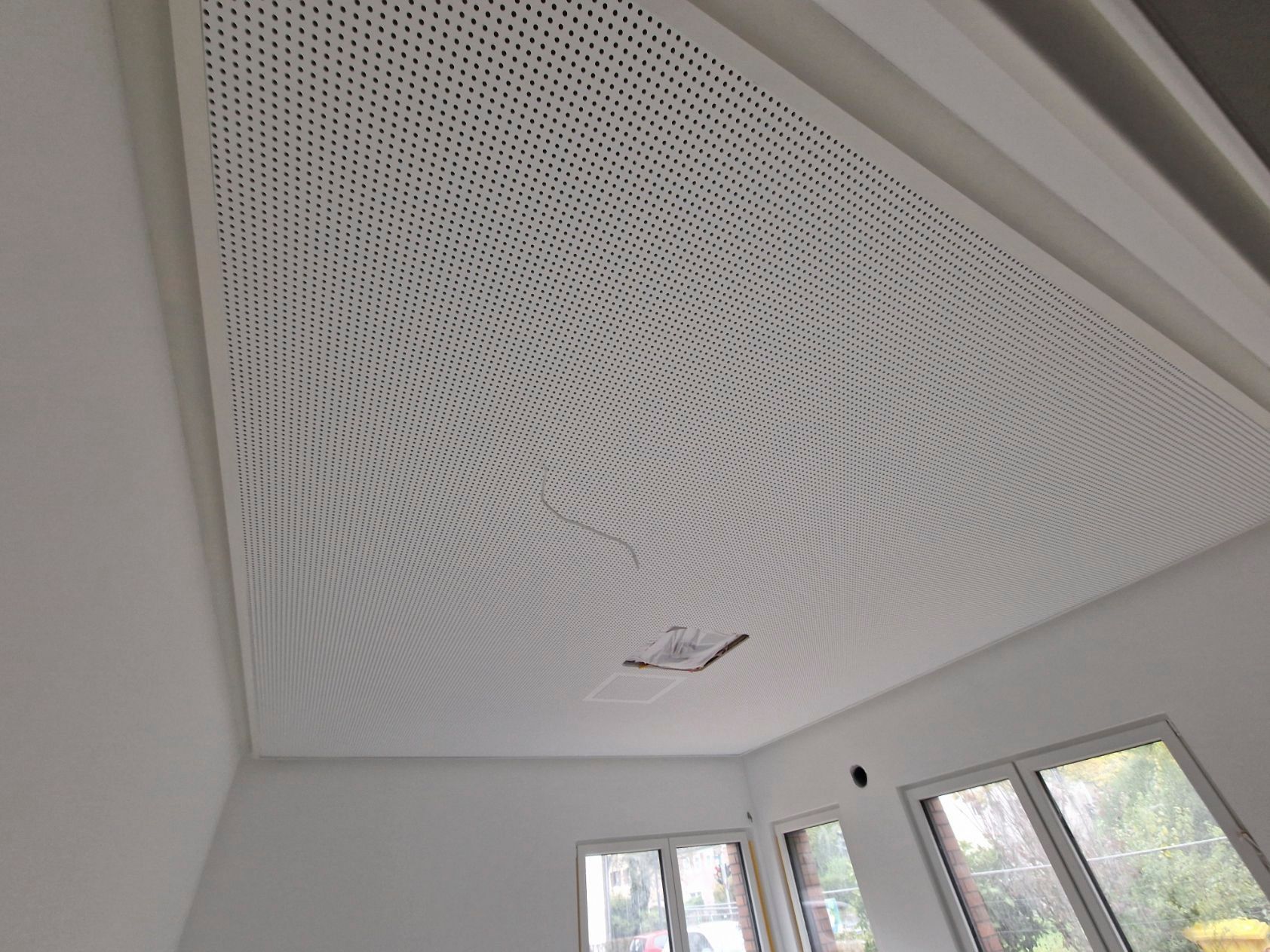 Weiße, perforierte Deckenplatte mit Wirbelmuster. Der Raum hat weiße Wände und ein Fenster.