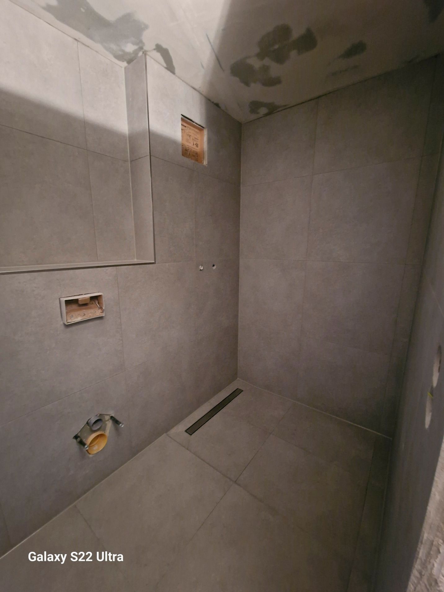 Grau geflieste Dusche im Bau; Abfluss, Sanitärarmaturen und Schaltkästen sind sichtbar.