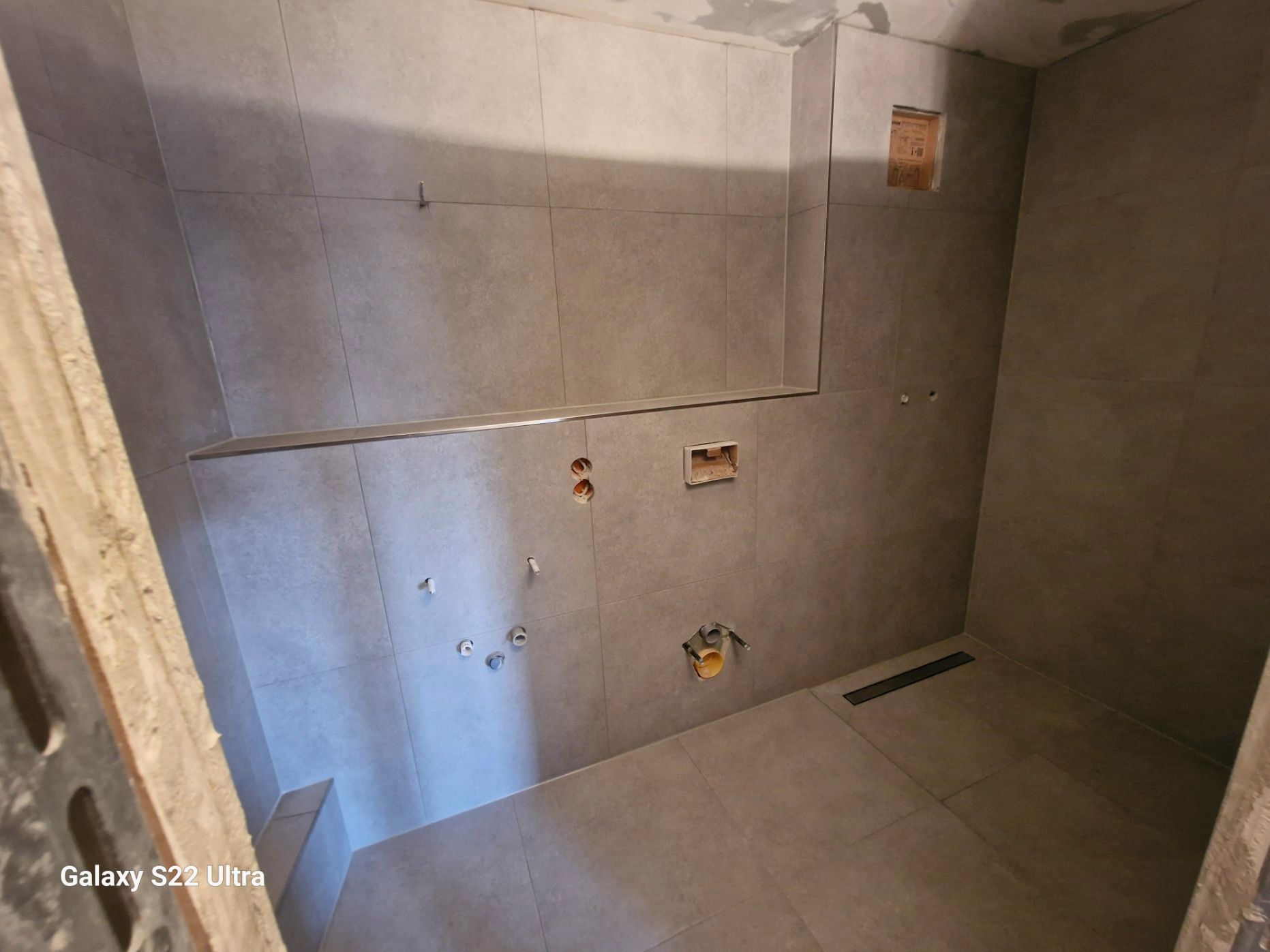 Innenansicht eines im Bau befindlichen gefliesten Badezimmers mit freiliegenden Elektro- und Sanitärarmaturen.