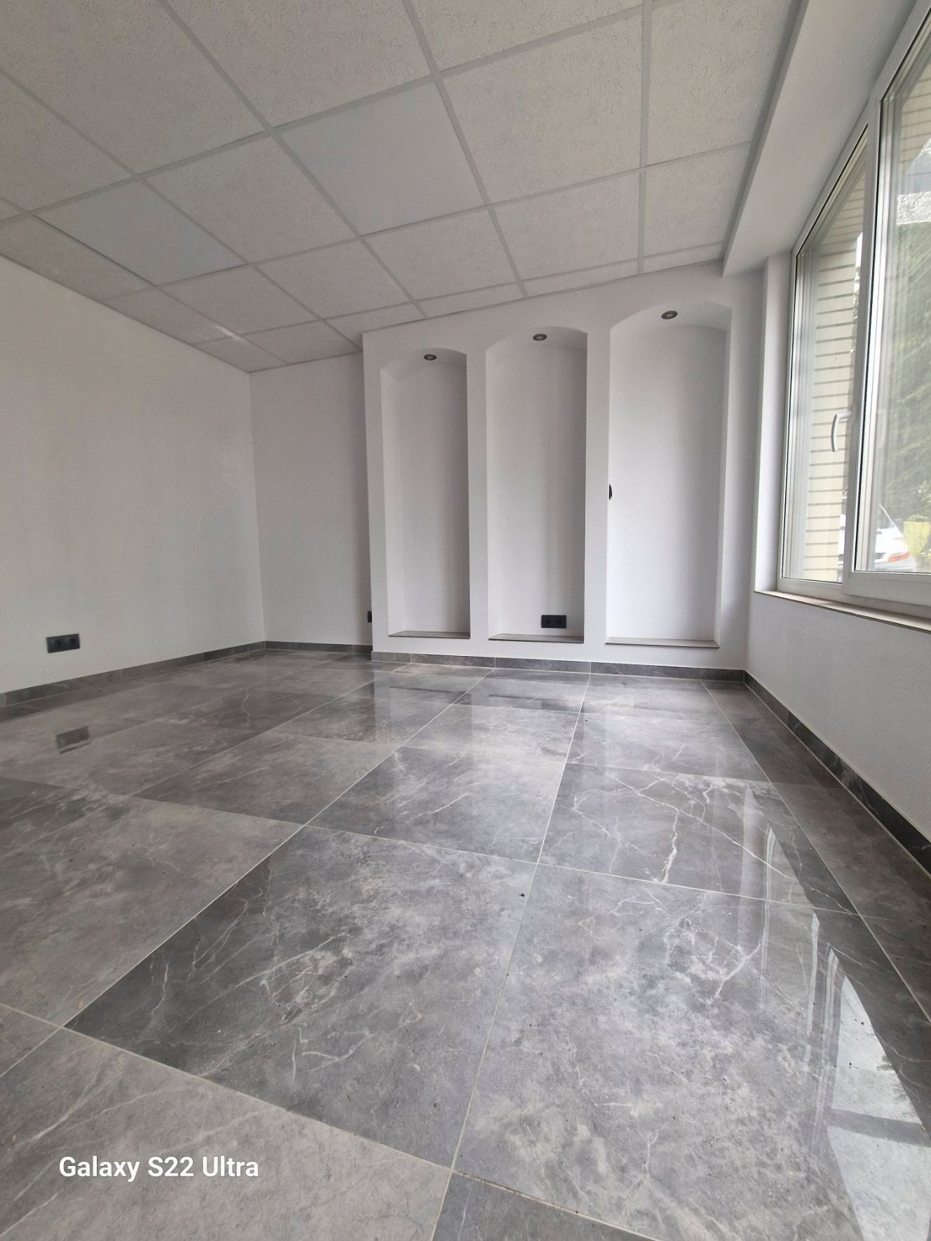 Leerer Raum mit grauem Marmorboden, weißen Wänden und Decke, drei gewölbten Wandnischen und einem Fenster.