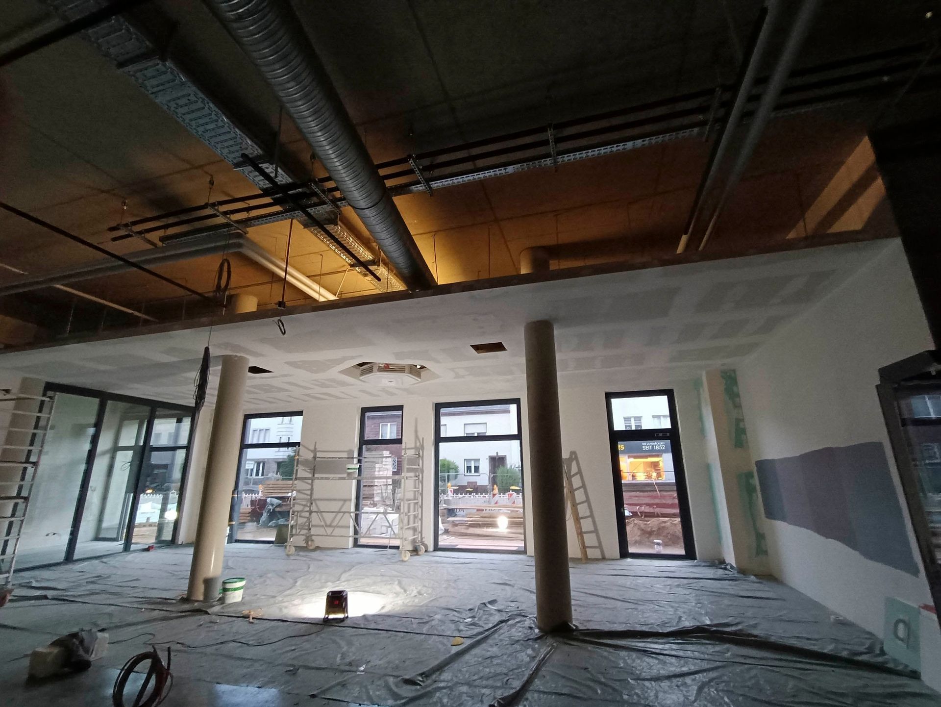 Innenansicht eines im Bau befindlichen Gebäudes mit offenen Fenstern, Trockenbauwänden, Betonpfeilern und freiliegender Deckeninfrastruktur.