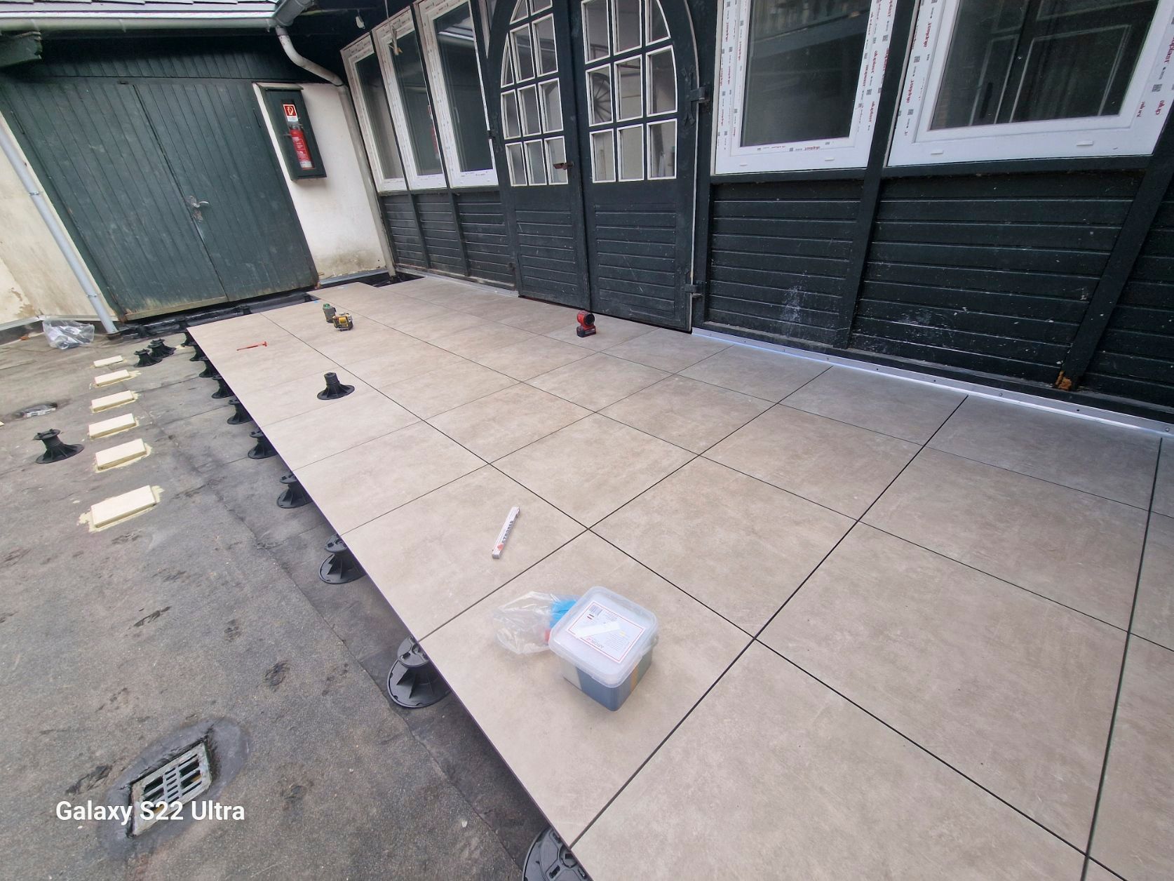 Außenterrasse mit beigefarbenen Fliesen. Schwarze Stützen und Gebäude.