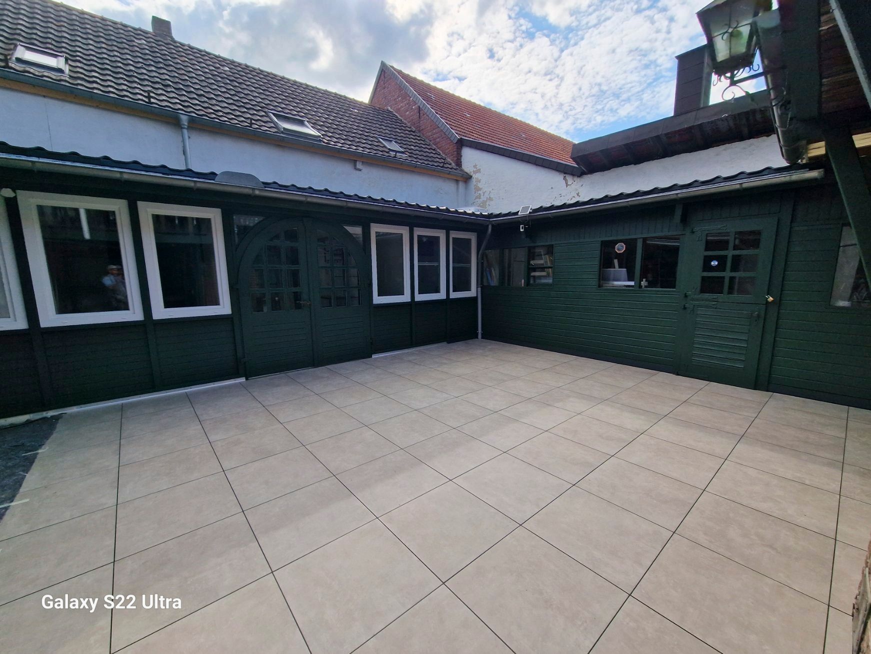 Grün gestrichenes Gebäude mit weiß gerahmten Fenstern, beige gefliester Terrasse und Blick auf einen bewölkten Himmel.