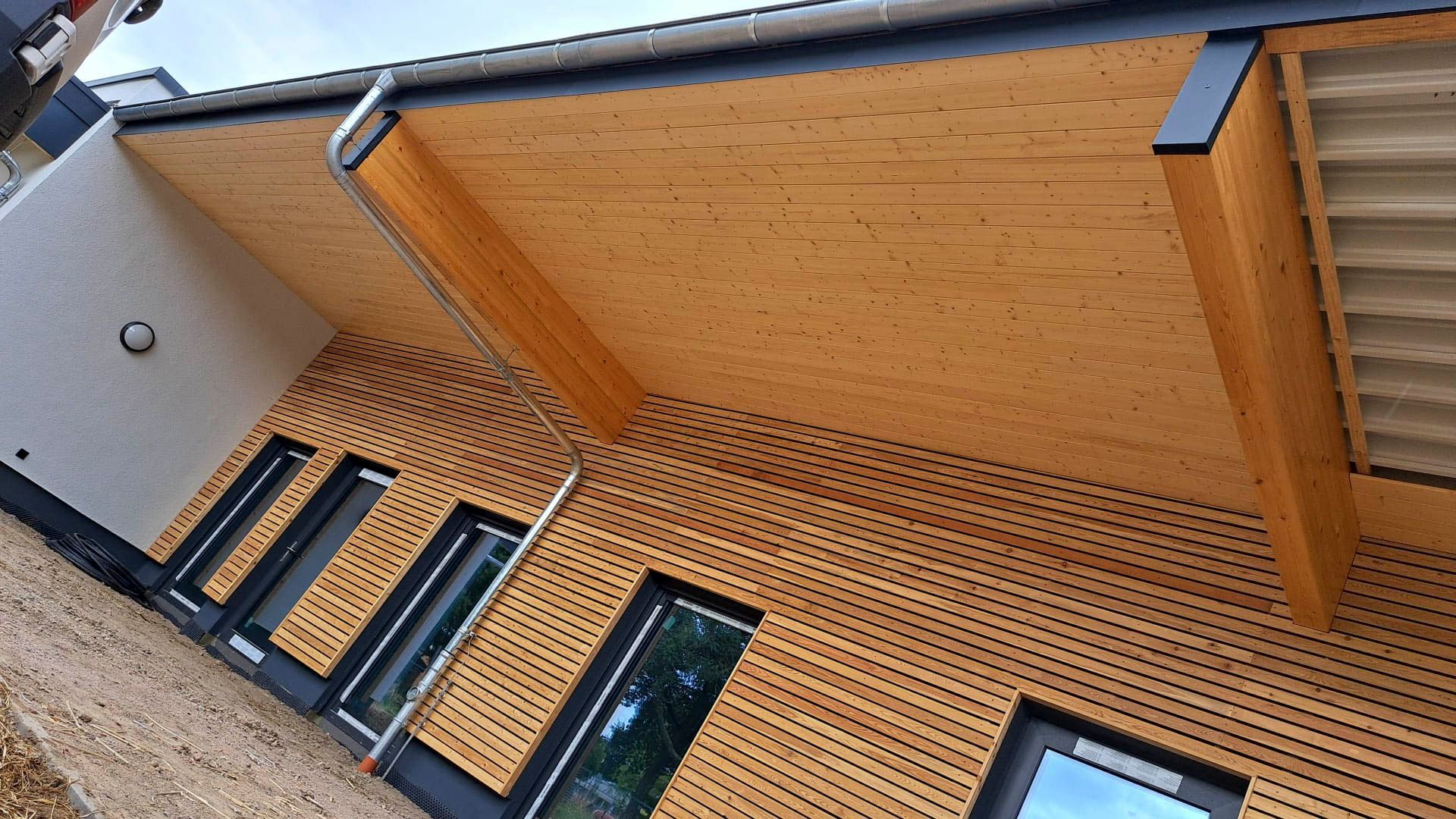 Außenansicht eines Gebäudes mit Holzverkleidung und Vordach. Schwarz gerahmte Fenster sind sichtbar.