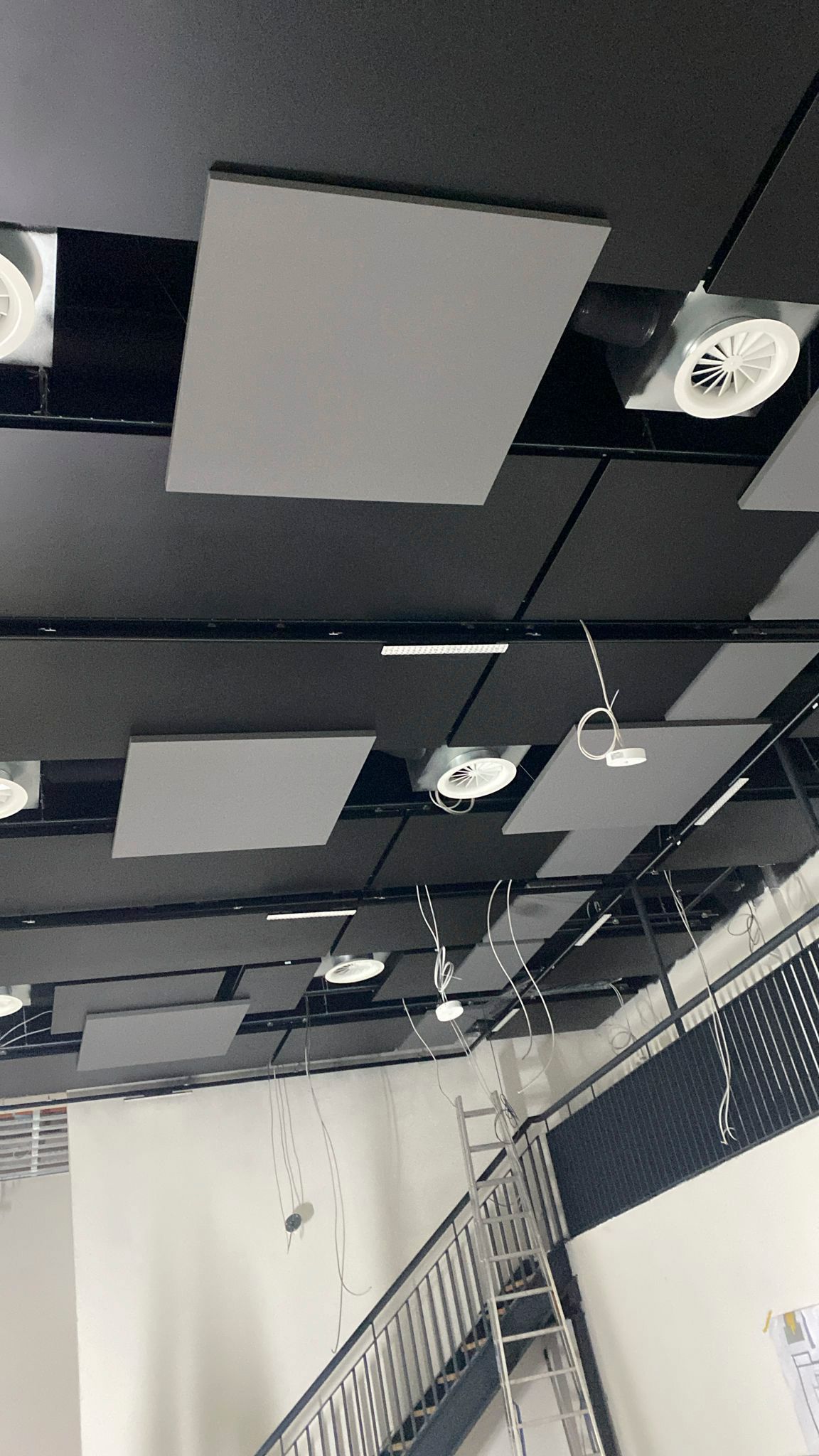 Graue Akustikplatten und runde Lüftungsschlitze an einer dunklen Decke; weiße Wände und darunter eine Metalltreppe.