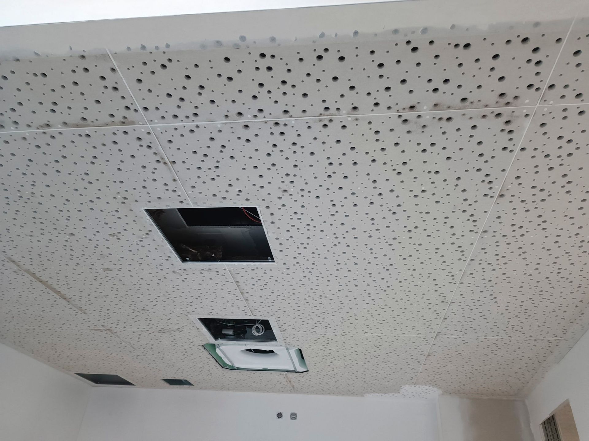 Weiße Deckenplatten mit schwarzen Perforationen, vertieften quadratischen Öffnungen zur Belüftung und einer Klimaanlage.